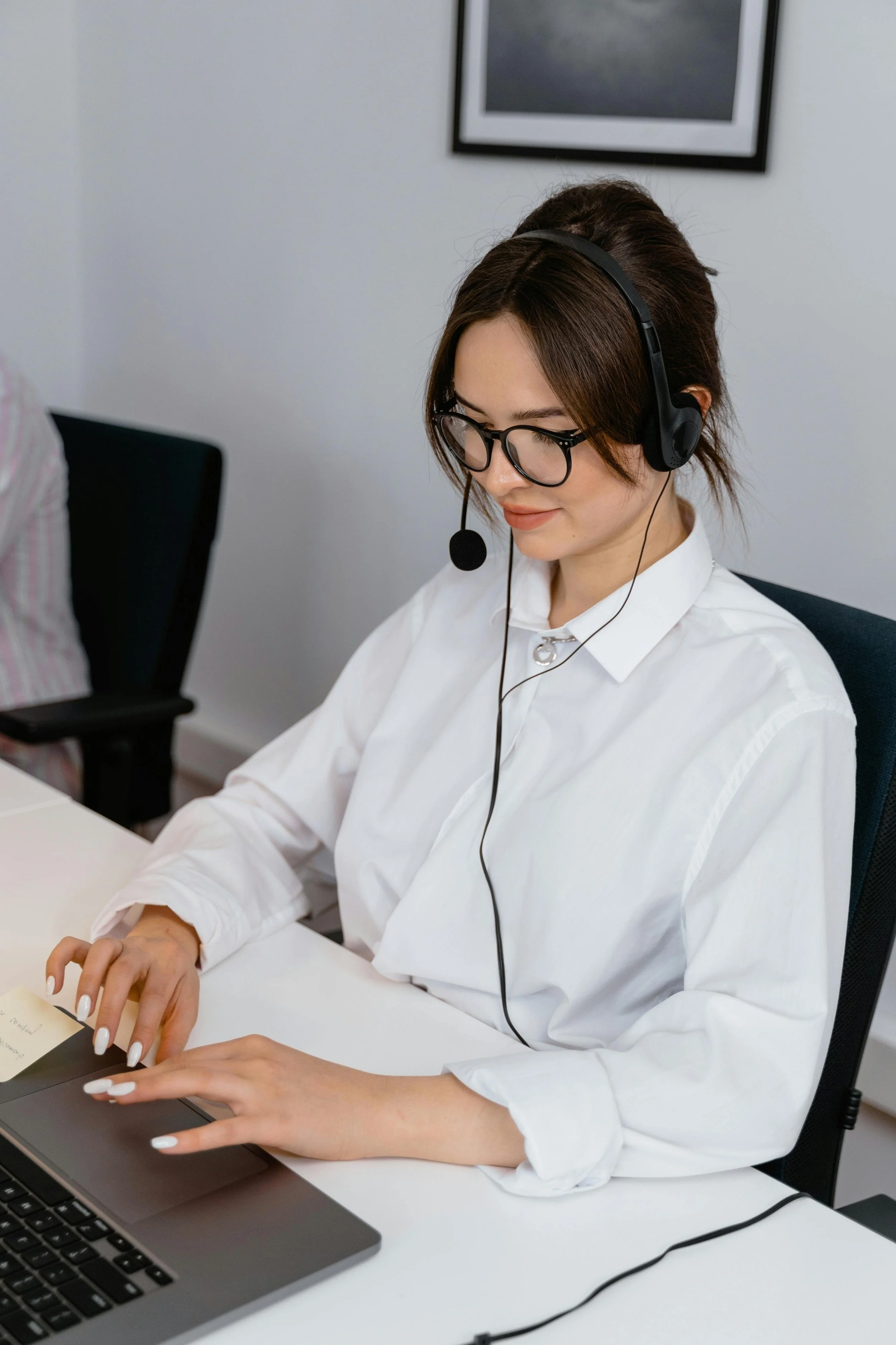A woman with glasses and brown hair, wearing a headset, white shirt, and a necklace, working on a laptop at a desk in an office.