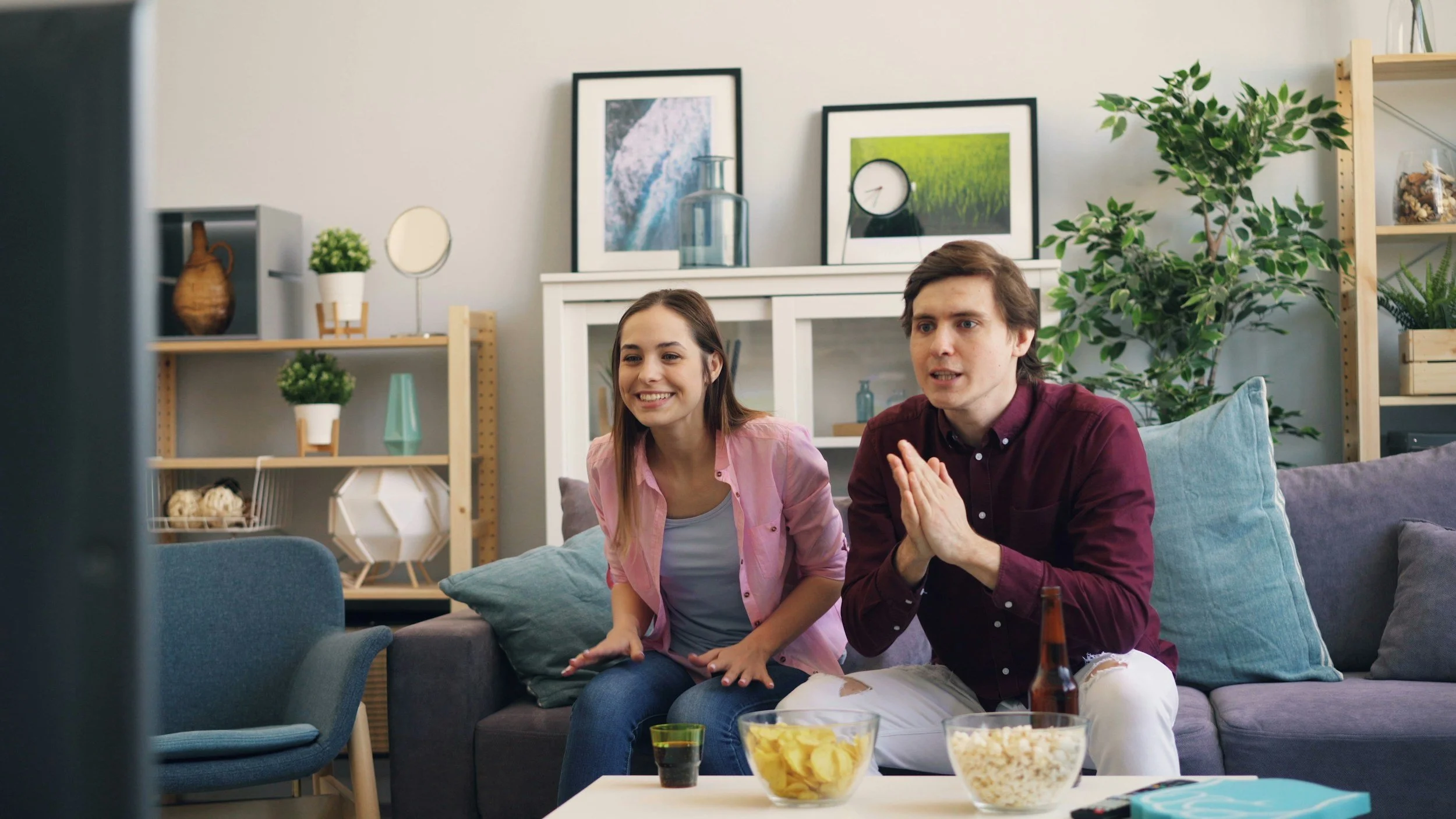 A young woman and a young man sitting on a gray couch, watching television in a cozy living room. The woman is smiling, while the man has his hands clasped in front of him. There are snacks and beverages on the table in front of them. The room is decorated with plants, framed pictures, and shelves with decorative items.