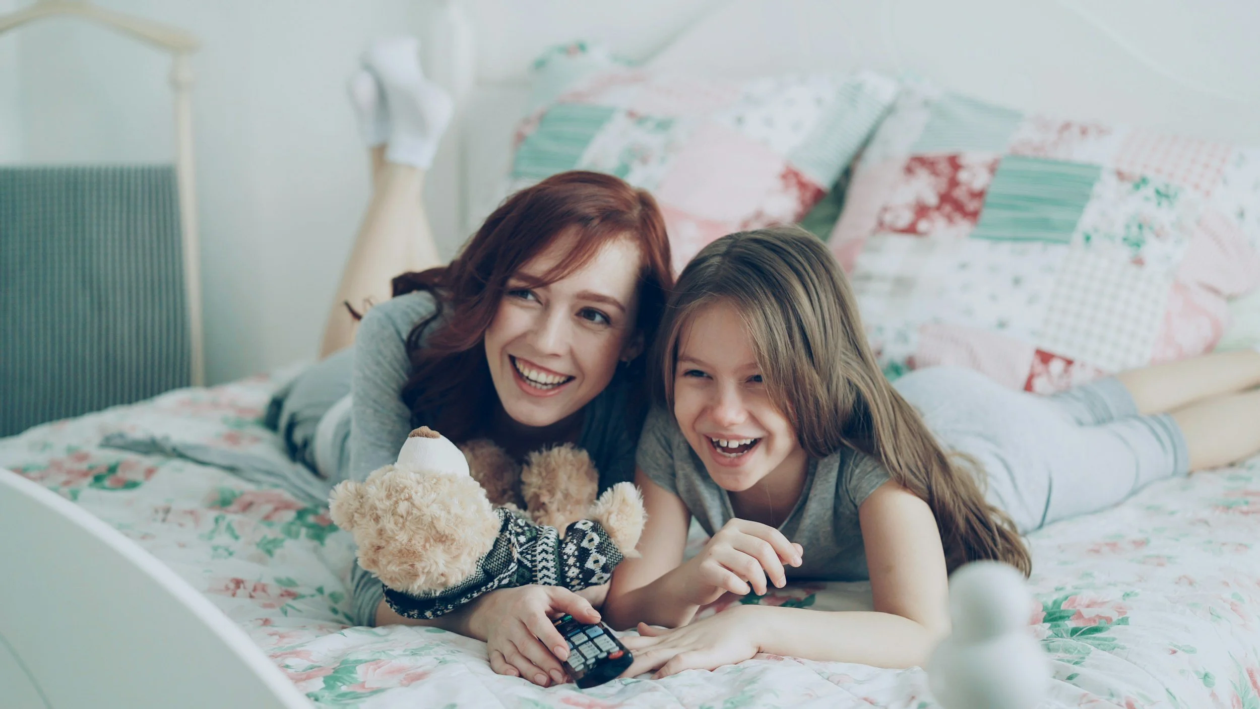 A woman and a girl lying on a bed, smiling and watching TV together, with a teddy bear in front of them.