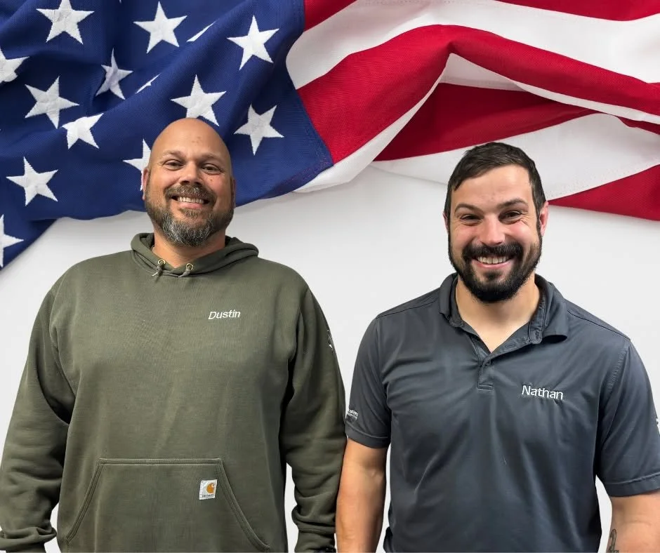 Two smiling men standing in front of an American flag backdrop. The man on the left is wearing a green hoodie with the name 'Dustin' on it, and the man on the right is wearing a dark gray polo shirt with the name 'Nathan' on it.