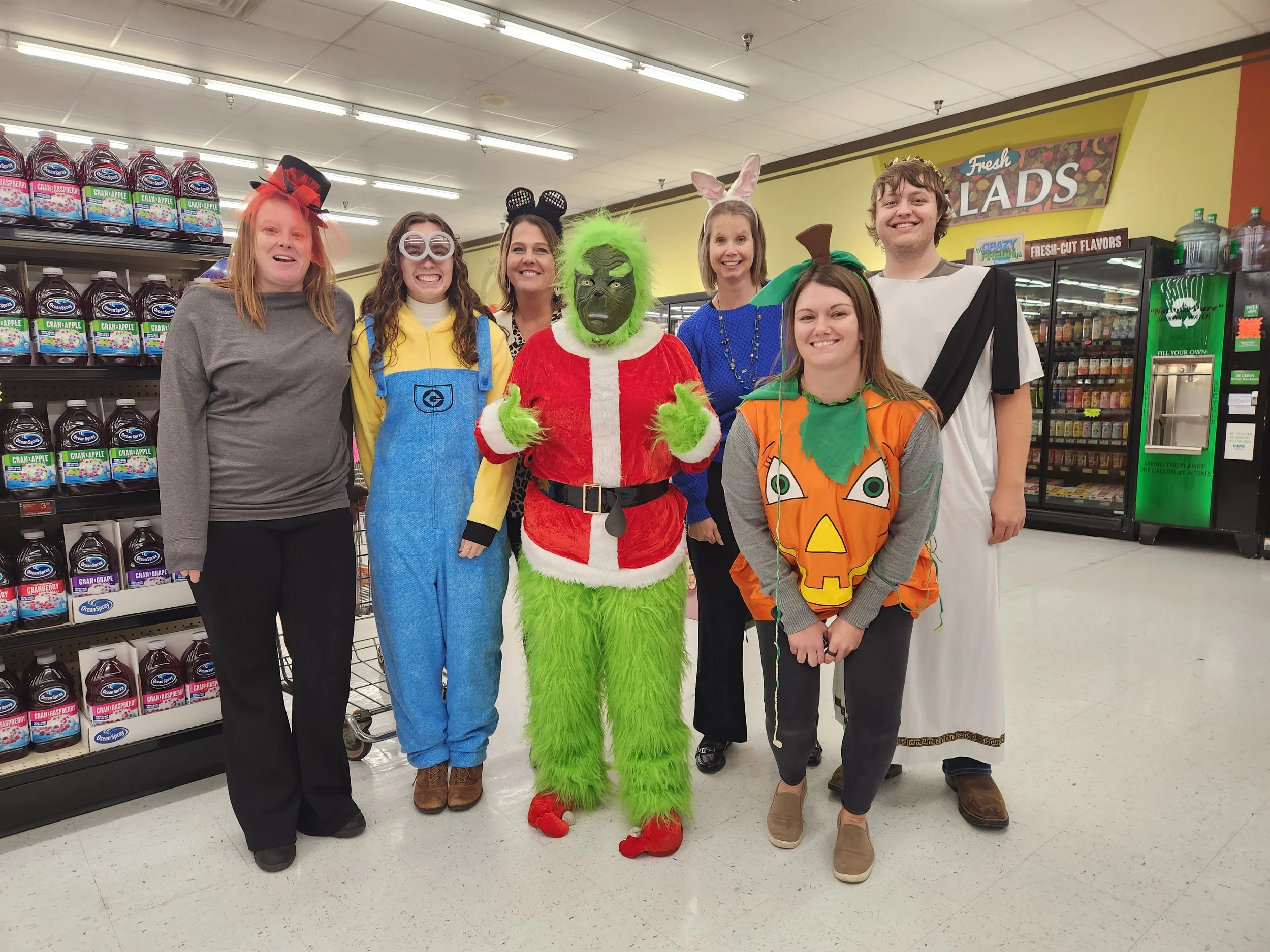 Group of six people dressed in costumes, standing in a grocery store aisle, with shelves of juice and candy behind them.