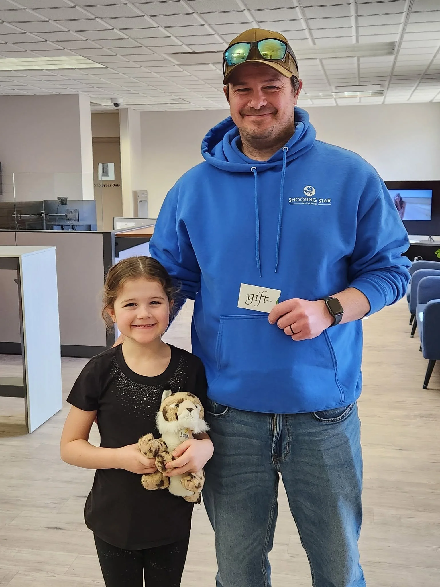 A man and young girl standing indoors, smiling at the camera. The girl is holding a stuffed tiger toy. The man is wearing a blue hoodie with a logo, sunglasses on his cap, and an Apple Watch. The girl is wearing a black shirt with a tiger face design.
