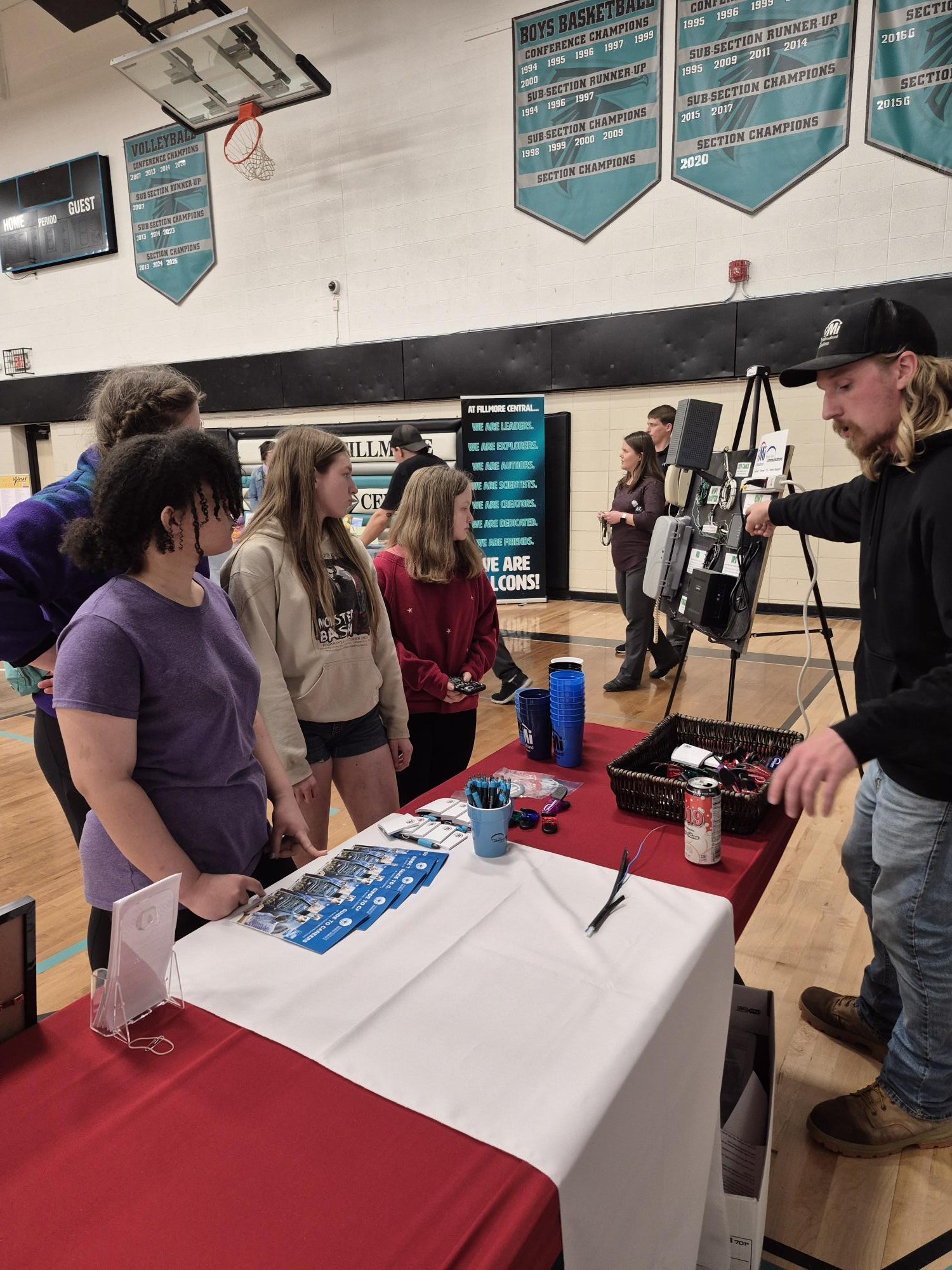 A group of people standing in line at a booth in a gymnasium with banners on the wall and a basketball hoop overhead. The booth has informational pamphlets, cups, and electronic devices on a table covered with a red tablecloth.