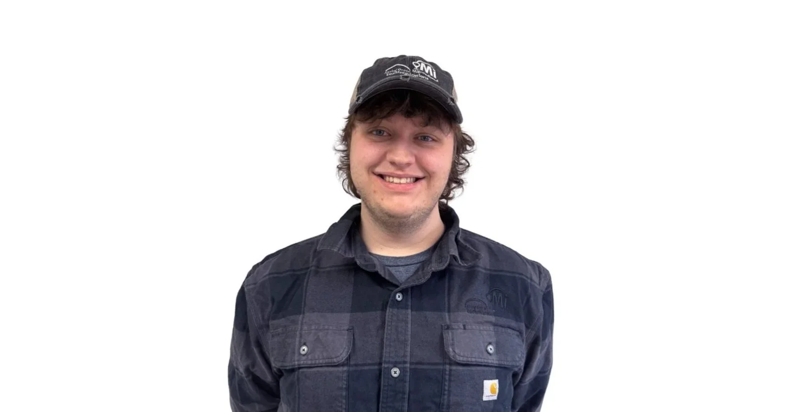 A young man with curly brown hair wearing a black cap and black Carhartt jacket, smiling against a white background.