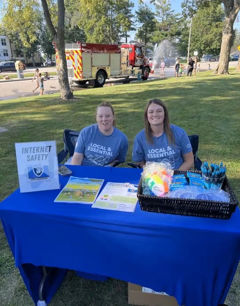 Two women sitting at a table with internet safety materials, outdoors in a park, with a fire truck spraying water in the background, and children playing.