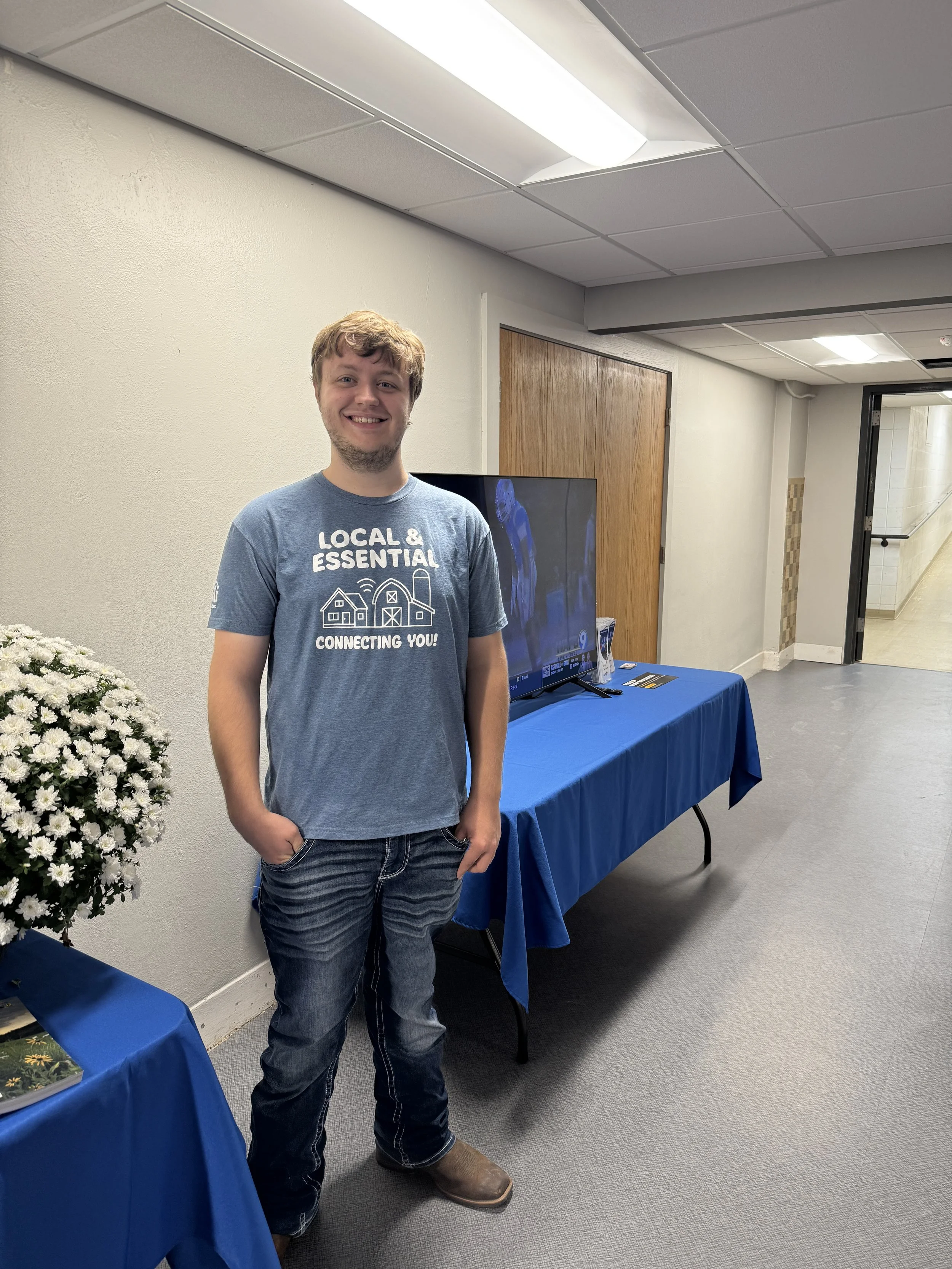 A young man with light brown hair and a light beard smiling while standing in a room with plain cream walls, a ceiling with fluorescent lights, and a hallway visible in the background. He is wearing a blue T-shirt with white text and graphics, dark jeans, and brown boots. There are blue-covered tables and chairs nearby, with a TV on a stand behind him and a potted plant with white flowers on the left.