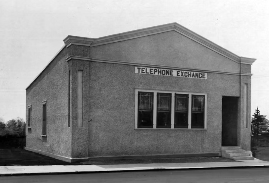 Black and white photo of a small building with a sign that reads 'Telephone Exchange' on the front.