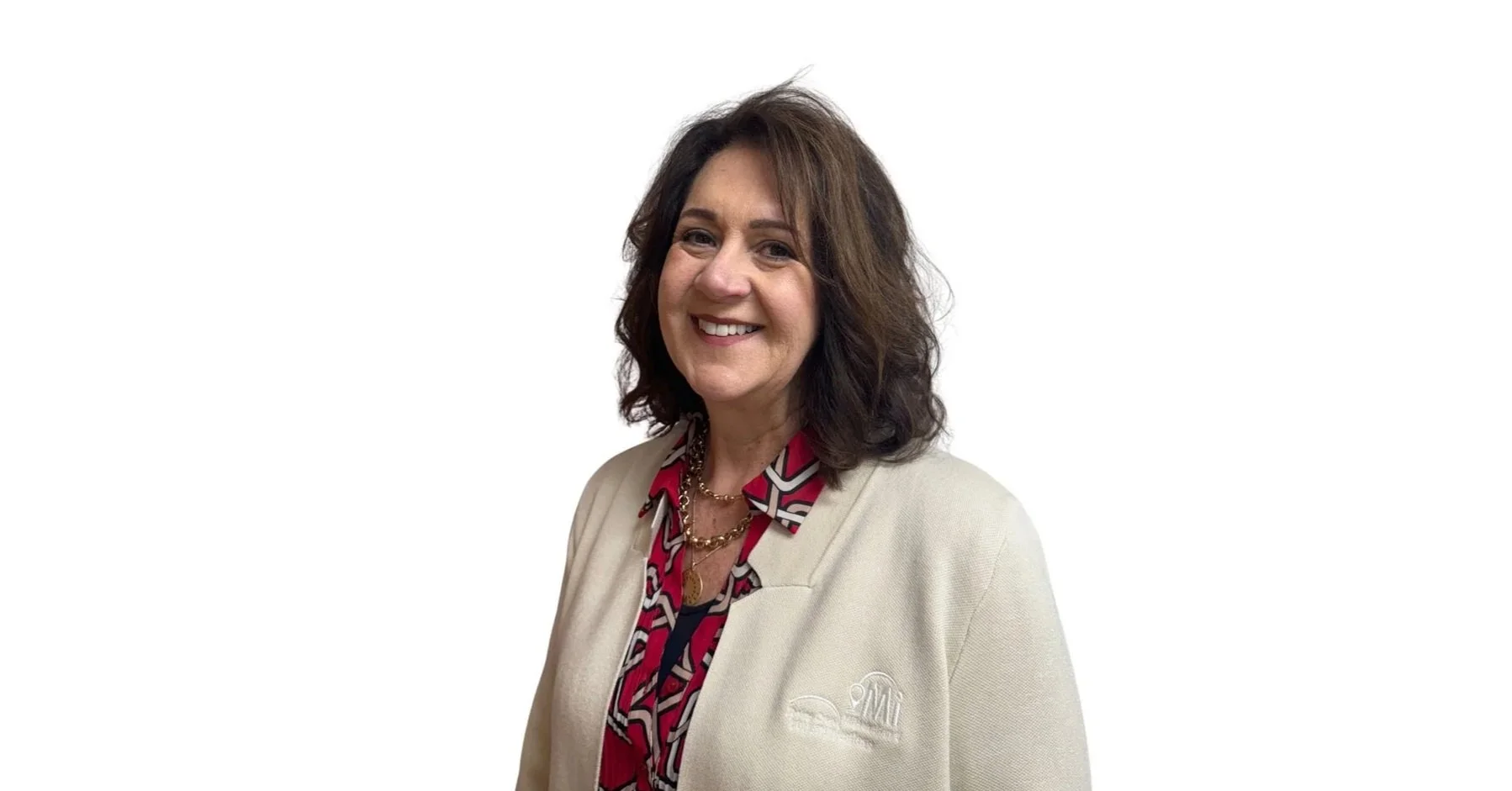A woman with mid-length dark brown hair, smiling, wearing a beige blazer over a red and black patterned blouse, standing against a plain white background.