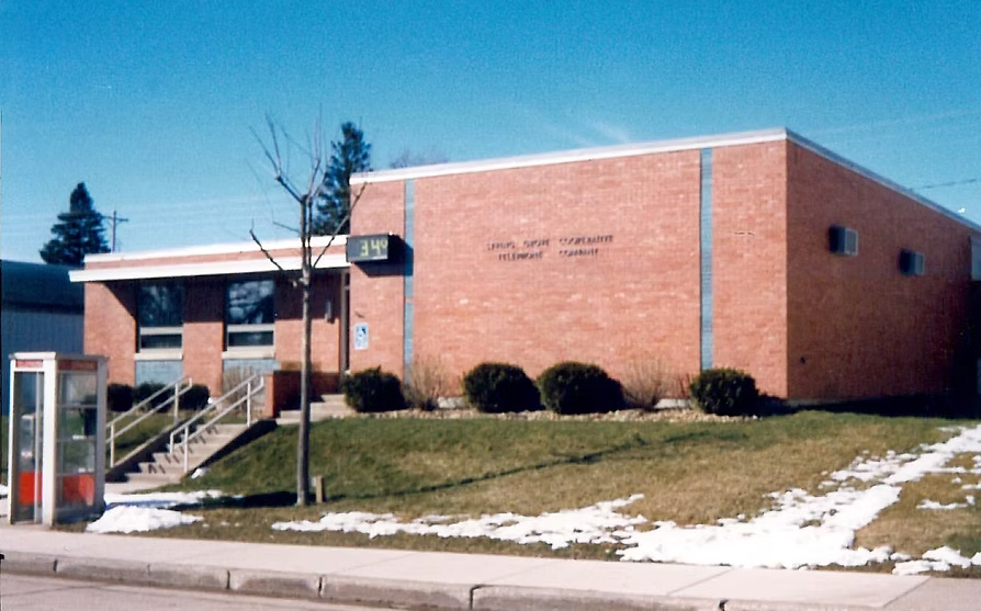A brick building with a sign indicating it is a Fire District Department, with steps leading up to the entrance, a bus stop, and patches of snow on the lawn.