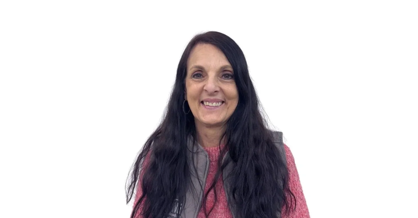 A woman with long dark wavy hair smiling, wearing a pink sweater and a gray vest, against a plain white background.