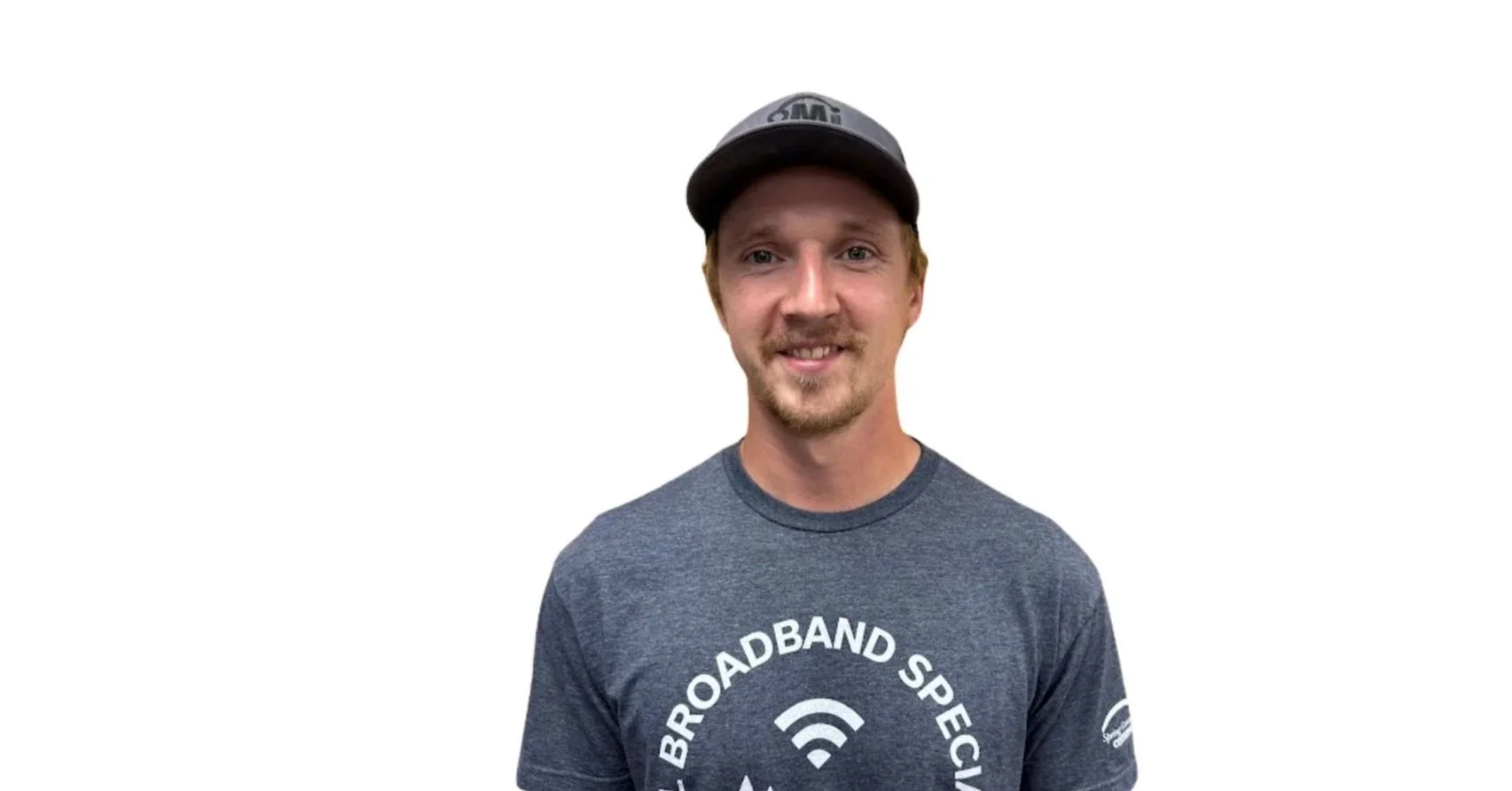 A young man with short red hair, a beard, and a mustache wearing a dark gray baseball cap and a gray T-shirt with a Wi-Fi icon and the text 'Broadband Specialist' around it, standing against a plain white background.