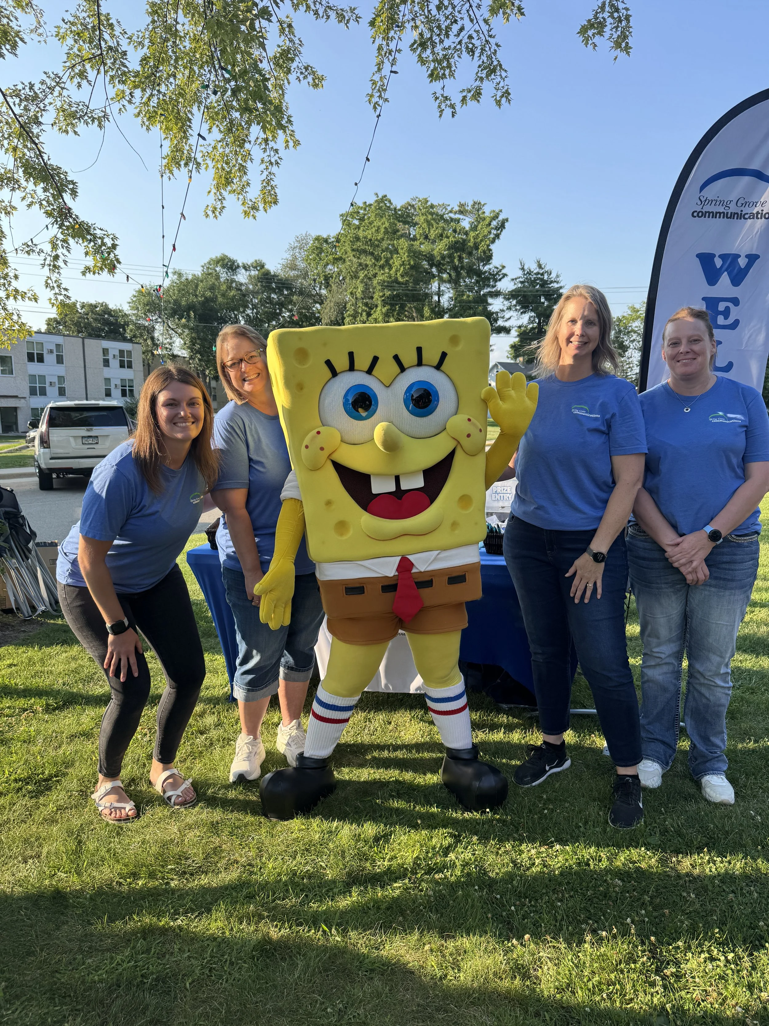 Four women standing outdoors on grass, posing with a person dressed as SpongeBob SquarePants. The women are smiling and wearing blue shirts, with trees and a flag in the background on a sunny day.