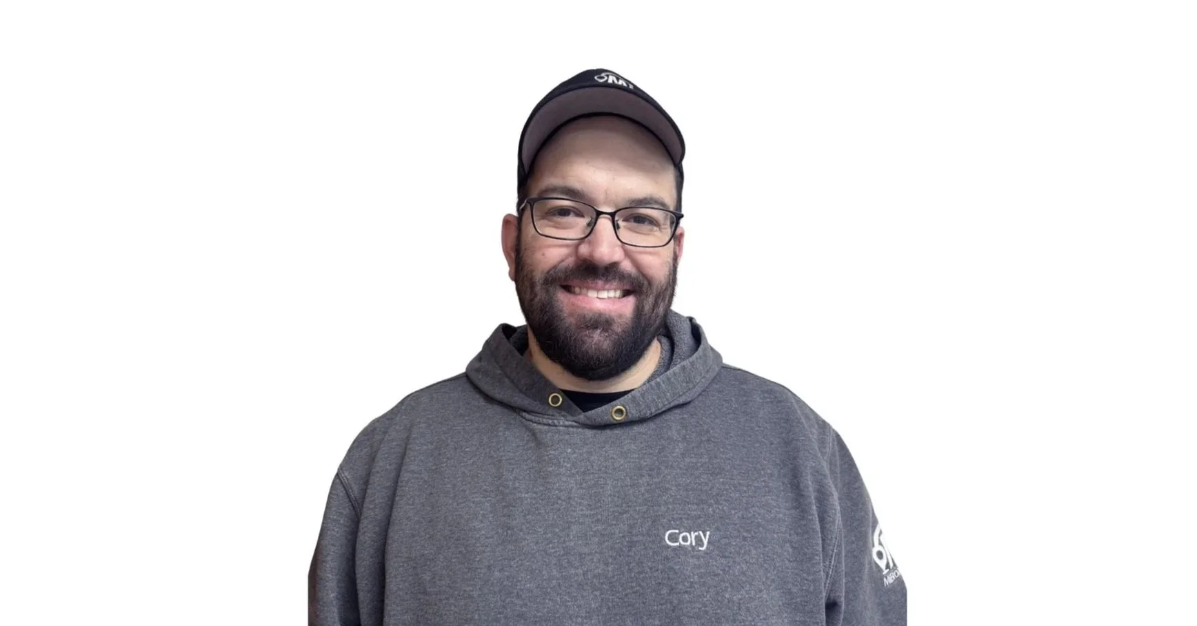 A man with glasses and a beard smiling, wearing a gray hoodie with "Cory" embroidered on it and a black cap, against a plain white background.