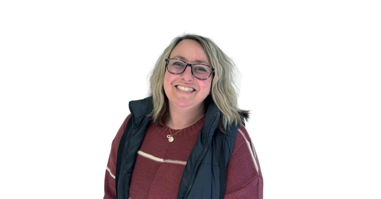A smiling woman with blonde, wavy hair, glasses, wearing a maroon sweater with white stripes, and a black vest, standing against a plain white background.