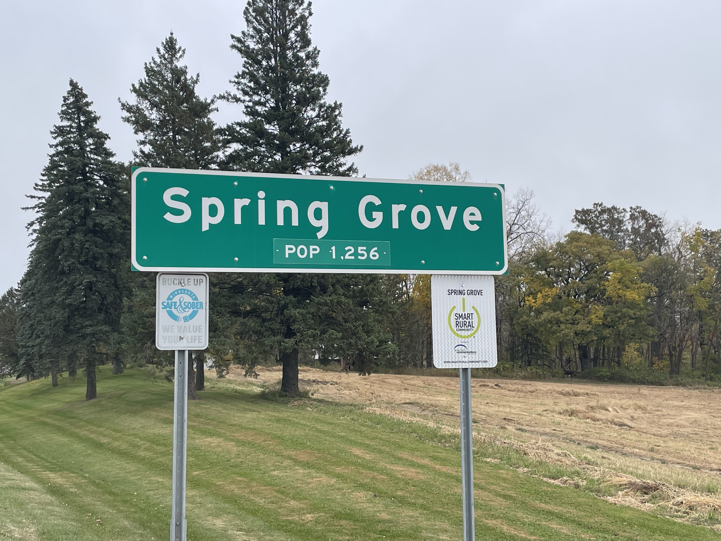 Green street sign reading 'Spring Grove' with population information, surrounded by trees and a partly cloudy sky.
