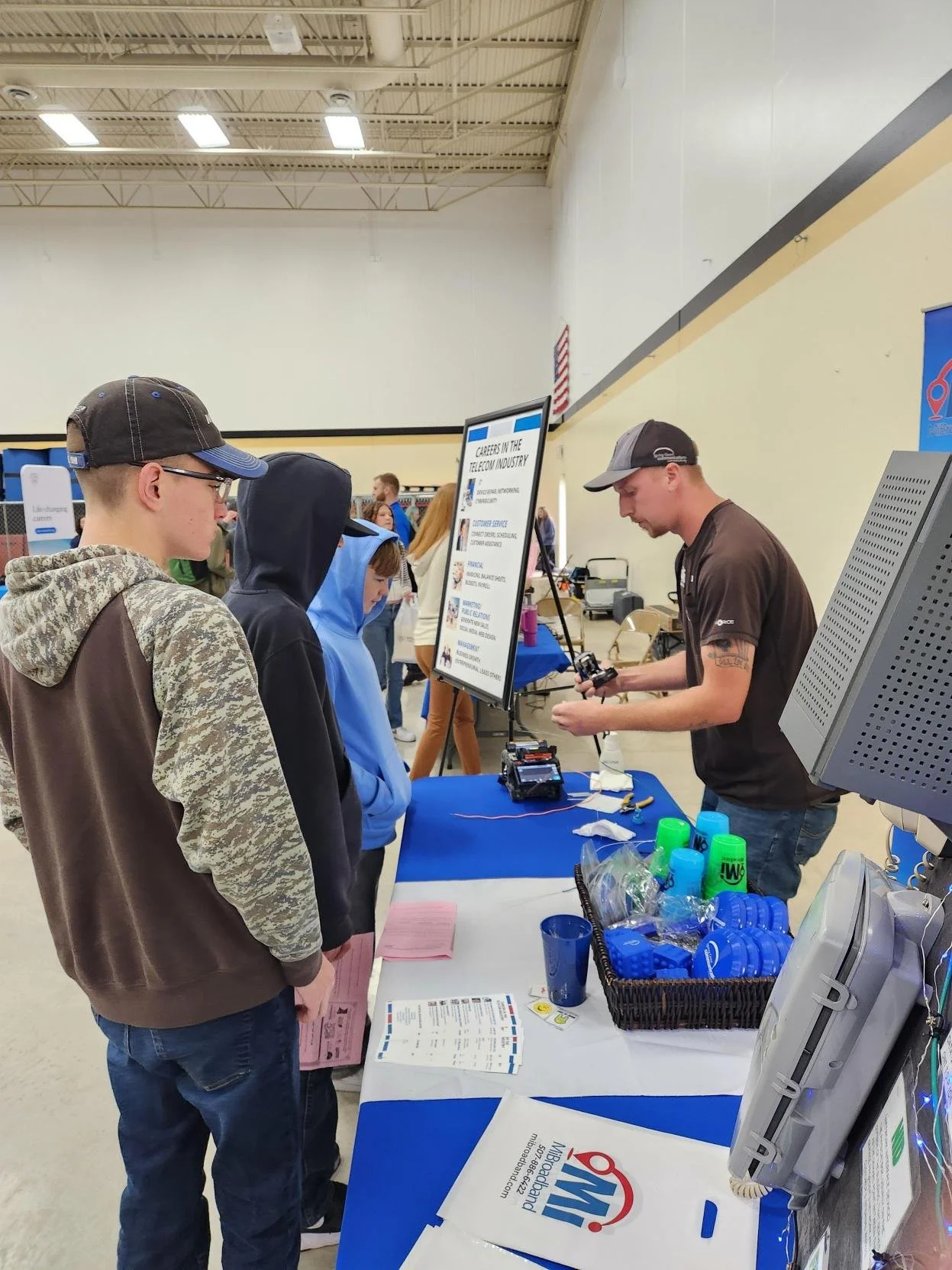 A man and young girl standing indoors, smiling at the camera. The girl is holding a stuffed tiger toy. The man is wearing a blue hoodie with a logo, sunglasses on his cap, and an Apple Watch. The girl is wearing a black shirt with a tiger face design.