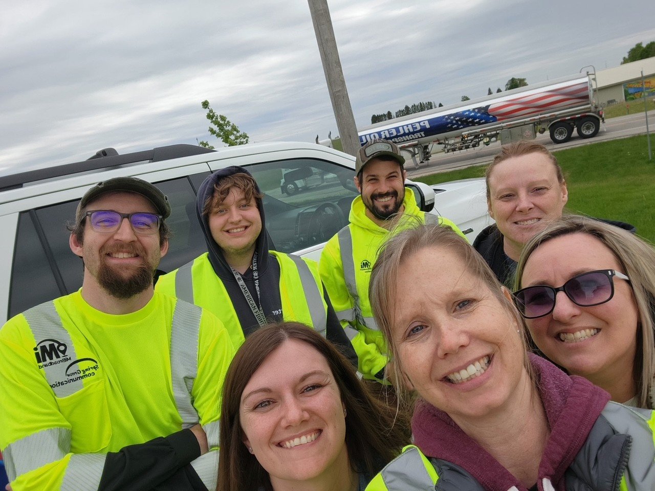Group of smiling people wearing yellow safety vests taking a selfie outdoors with a white vehicle and a trailer with an American flag design in the background.