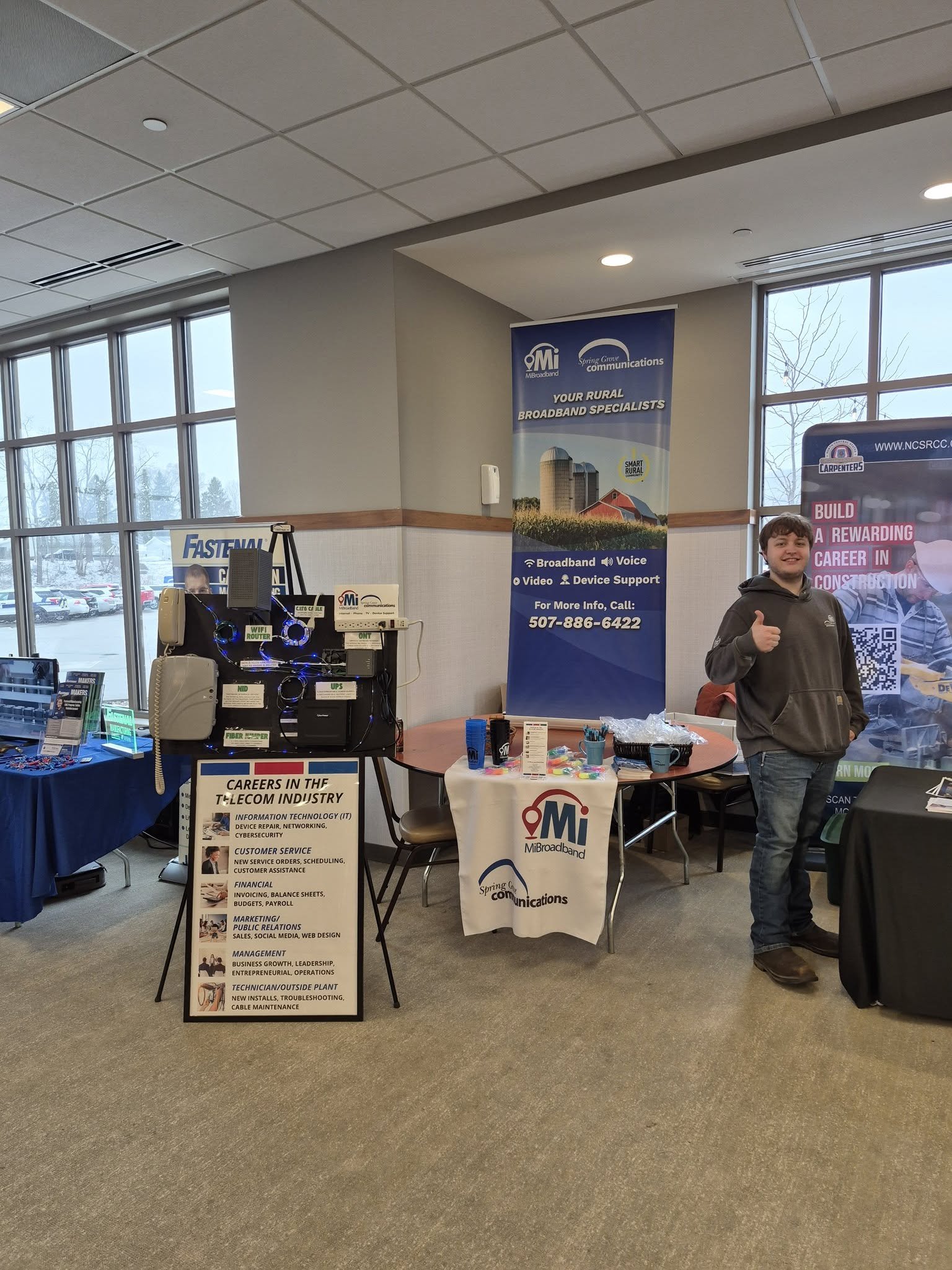 A man standing at a booth promoting broadband and telecommunications services at an indoor event. The booth has banners and informational signs, and the man is smiling and giving a thumbs-up.
