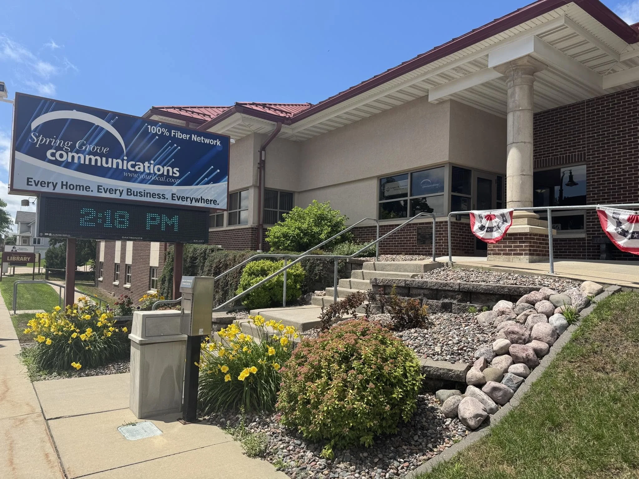 Exterior of a building with a large sign that reads 'Spring Grove Communications, 100% Fiber Network,' and a slogan underneath stating 'Every Home. Every Business. Everywhere.' There is a sidewalk with yellow flowers and landscaping, a set of steps leading to the entrance, and patriotic bunting decorations. The sky is clear and blue.