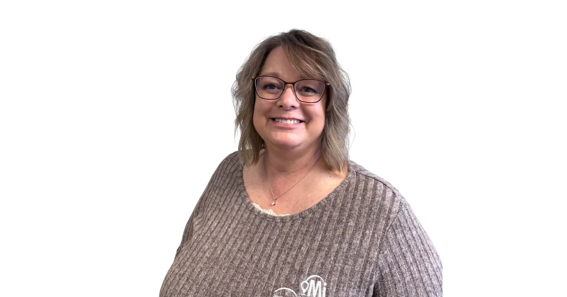 A woman with shoulder-length wavy gray hair and glasses smiling at the camera, wearing a brown knit sweater with a monogrammed logo on the front.