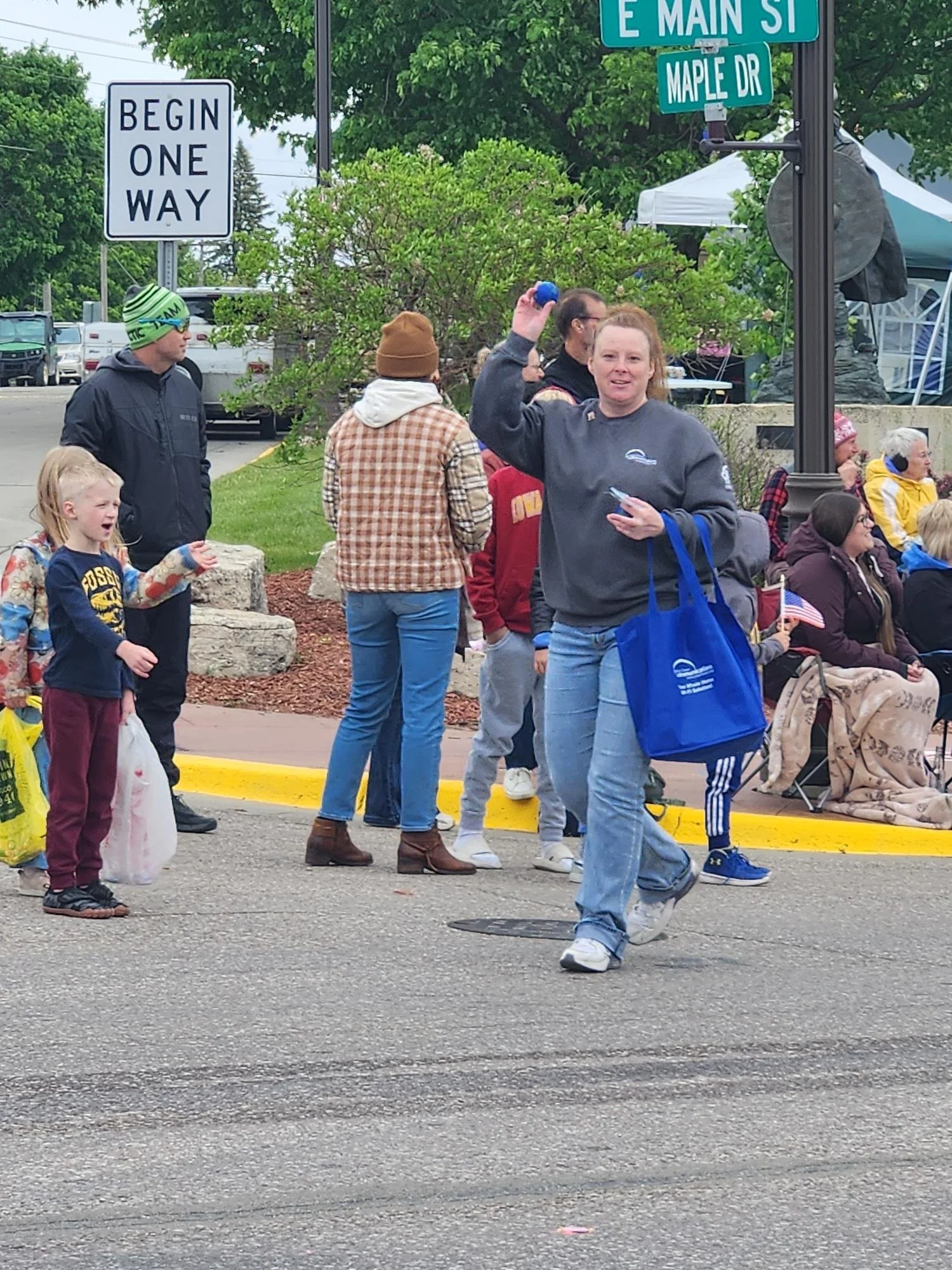 A group of people gathered outdoors on a city street, some sitting on benches and others standing. The scene includes children, adults, street signs, and a man waving with a smile, holding a small object in his hand. There are cars parked in the background and a tent set up nearby.