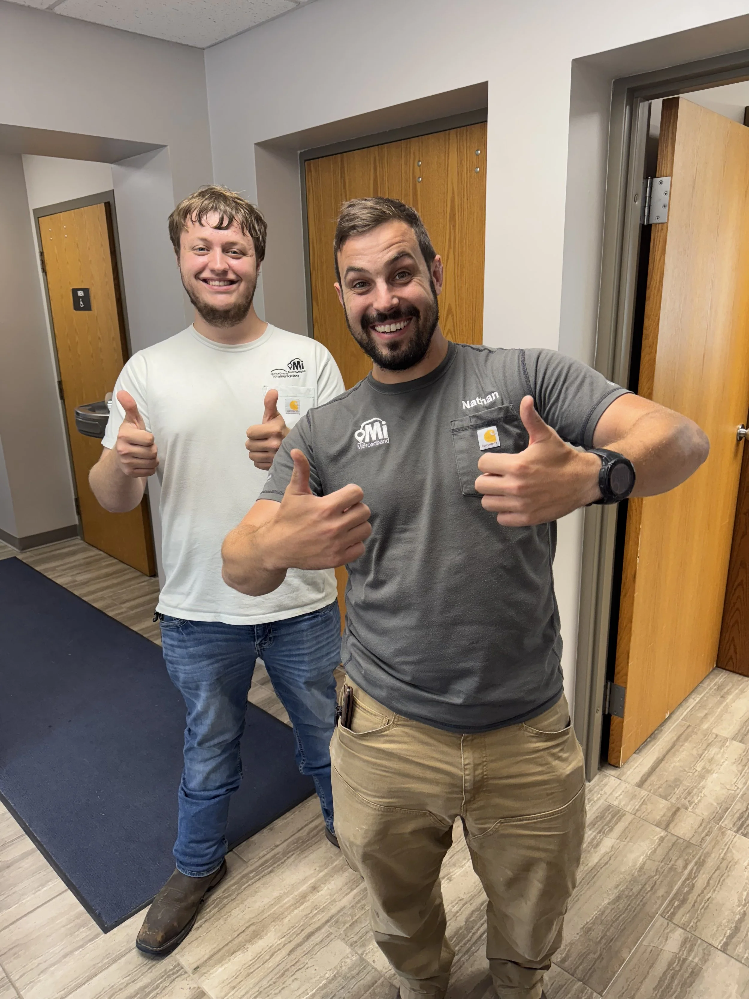 Two smiling men giving thumbs up inside a building, standing on a wood floor near restroom doors, one wearing a gray shirt with the name 'Nathan' and the other in a white shirt.