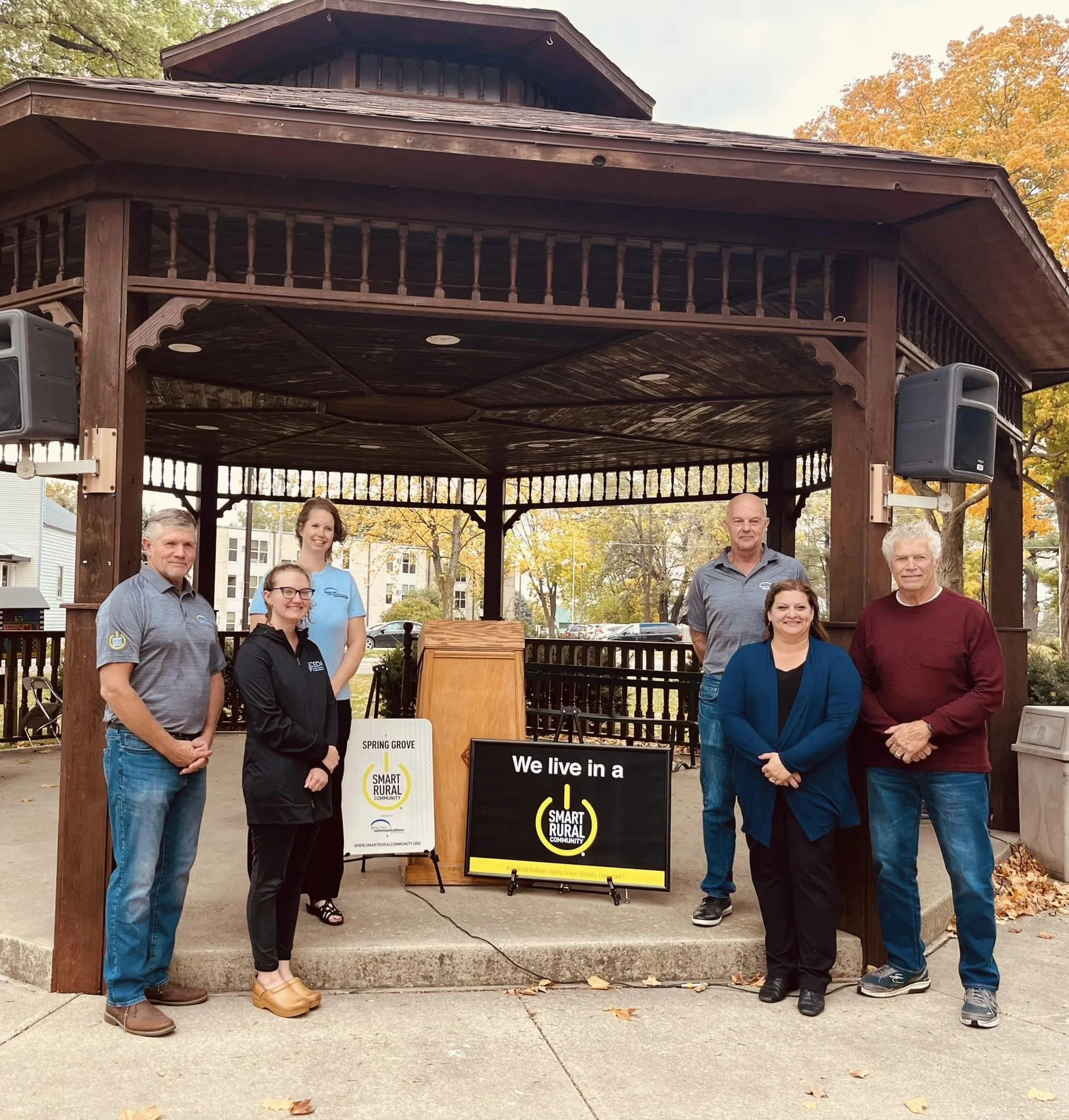 Group of people standing under a wooden gazebo with signs that say 'Smart Rural Community' and 'We live in a Smart Rural Community' in a park with autumn leaves on trees in the background.