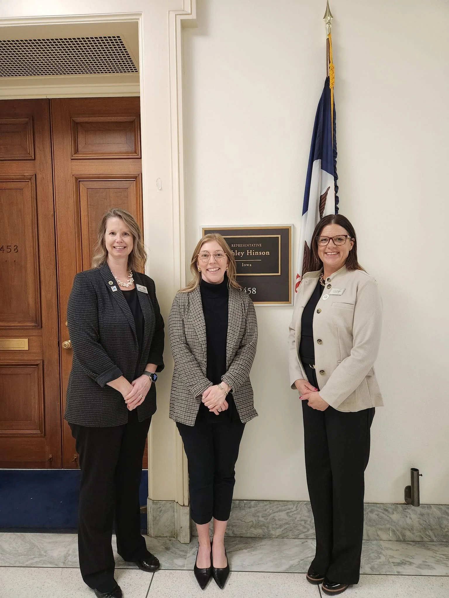 Three women standing in front of a door with a nameplate that reads 'Representative Ashely Hinson Iowa' and a flag beside the door. The women are smiling and dressed in professional attire.