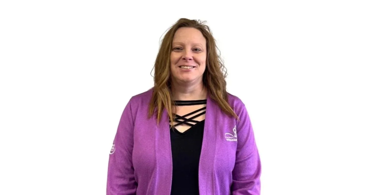 A woman with long, wavy, reddish-brown hair, wearing a purple jacket over a black top with crisscrossing straps, smiling and standing against a plain white background.