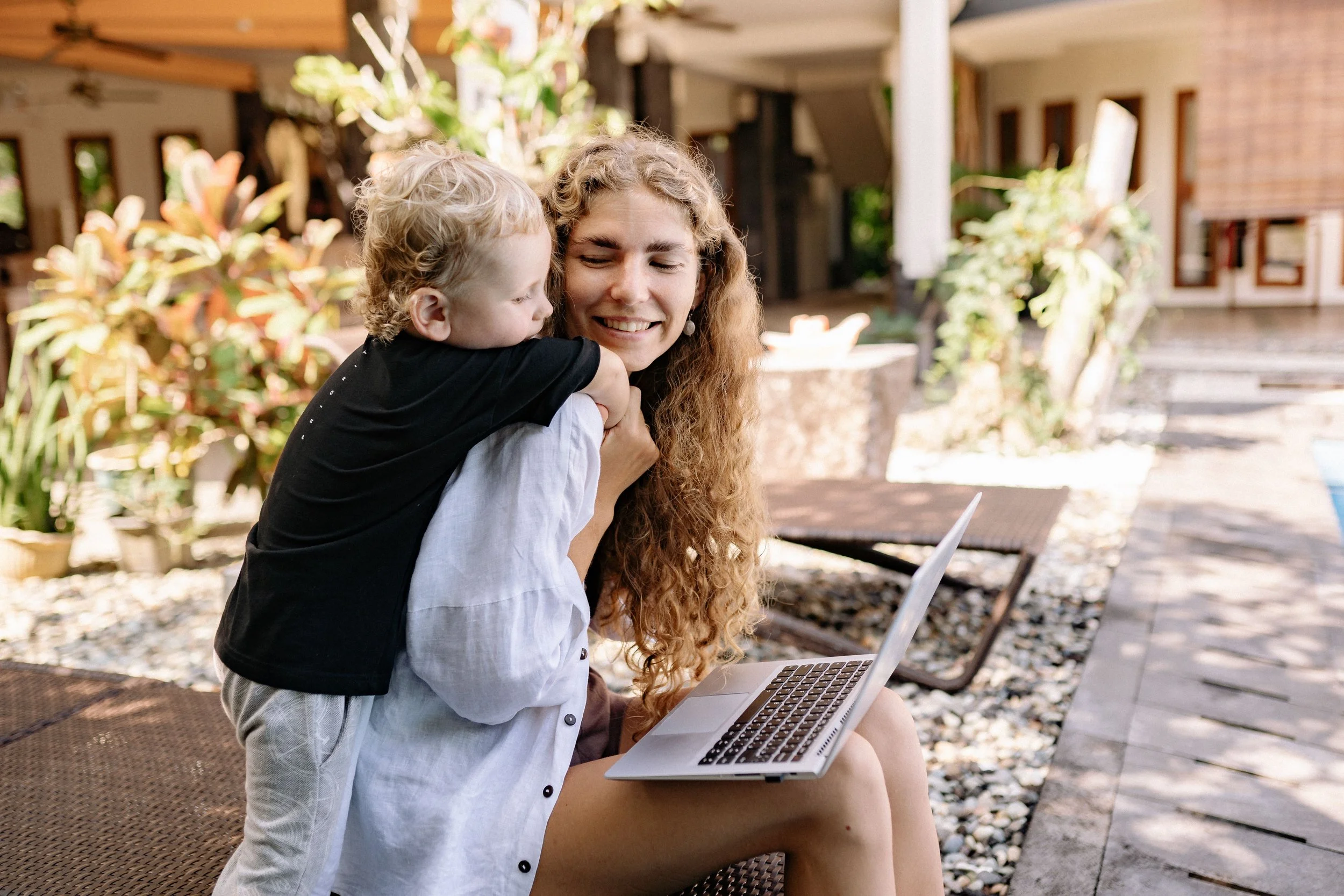 A young woman sitting outdoors on a bench holding a laptop on her lap, smiling, with a young boy hugging her from the side, both enjoying a moment together in a garden or backyard setting with plants and buildings in the background.