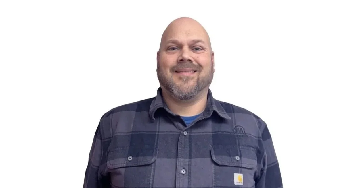 A bald man with a beard smiling, wearing a dark plaid shirt, standing against a white background.