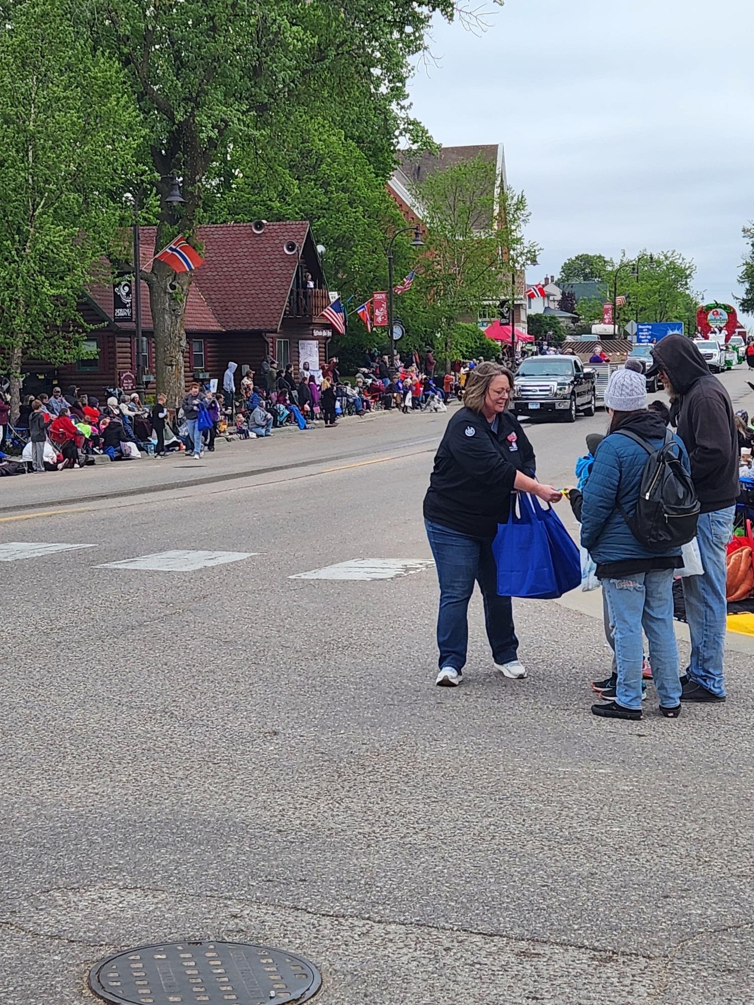 A street scene with people waiting in line along the sidewalk, some seated on benches and others standing. The street is decorated with flags and banners, and a large building with a peaked roof is visible in the background. The scene appears to be part of a parade or public event.