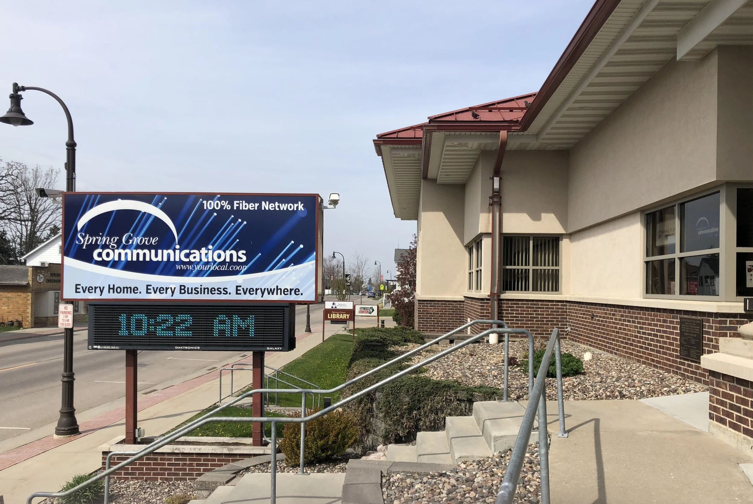 A sign advertising Spring Grove Communications with a blue background and white text, stating "100% Fiber Network" and "Every Home. Every Business. Everywhere." next to a building with a brick and beige exterior, a red roof, and a sidewalk with steps and a metal railing.