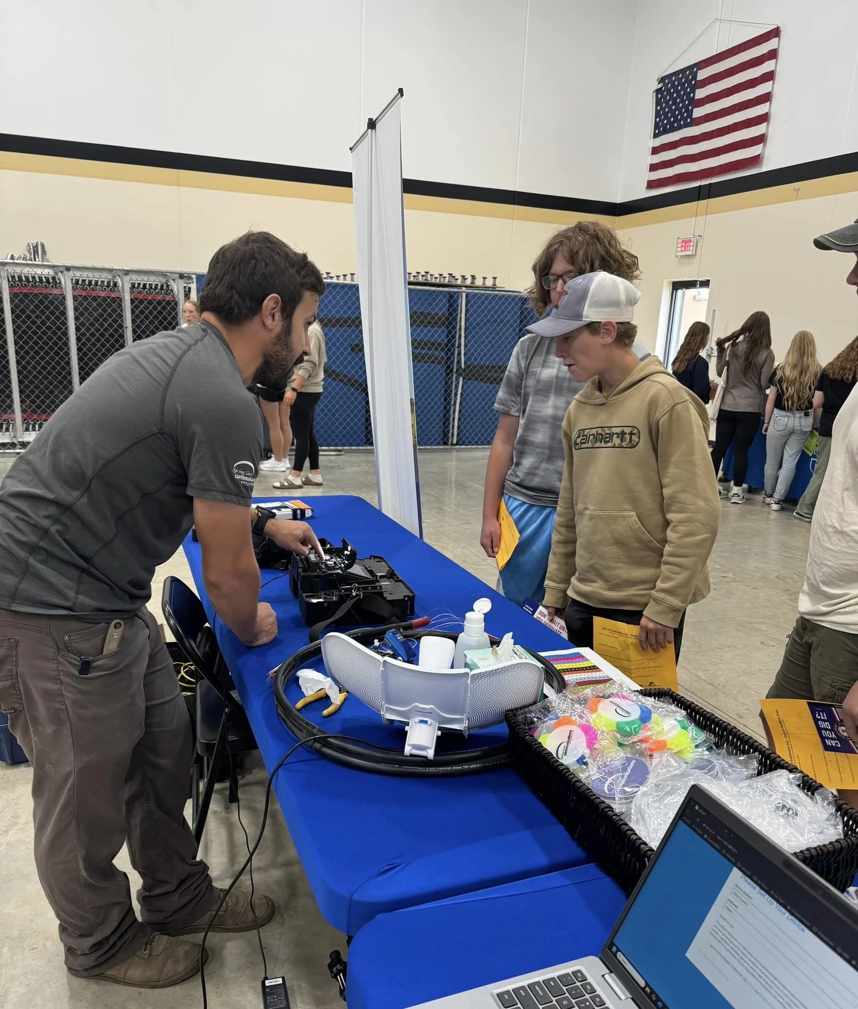 People are gathered around a table with electronic devices and equipment, engaging in a demonstration or discussion at an indoor event with an American flag hanging on the wall in the background.