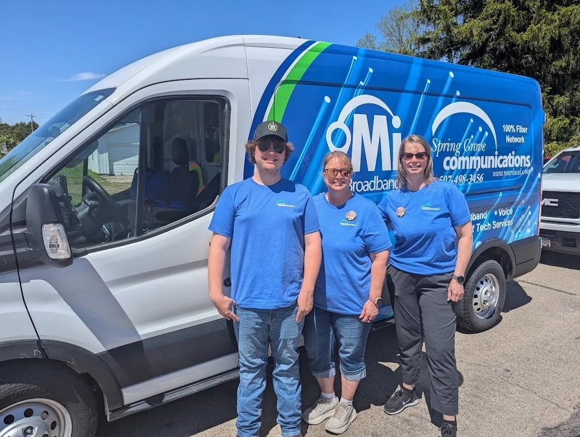 Three people standing in front of a Spring Grove Communications van, all wearing blue shirts and smiling.