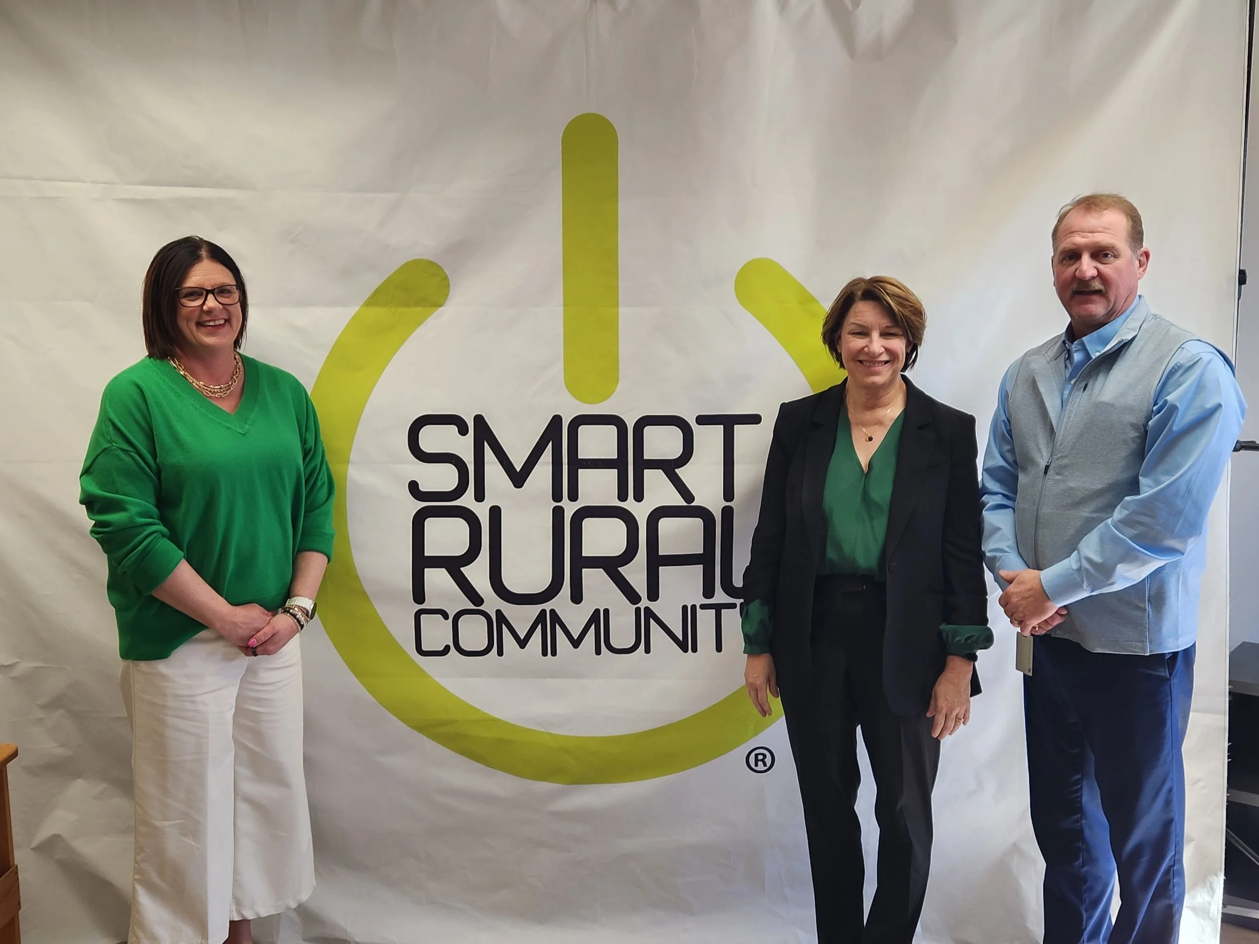 Four people standing in front of a white background with the words "Smart Rural Communities" written on it. The group includes three women and one man, all smiling and dressed in business casual attire.