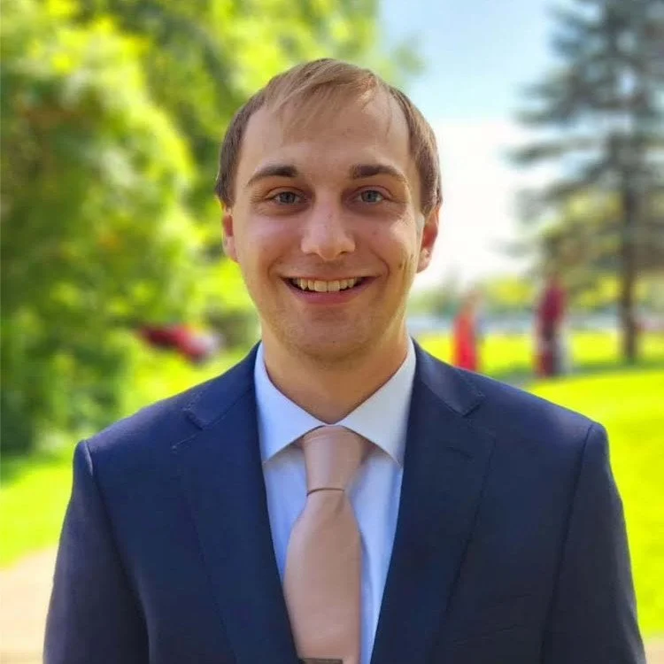 A young man in a navy suit, white shirt, and beige tie outdoors, smiling with a background of trees and grass.