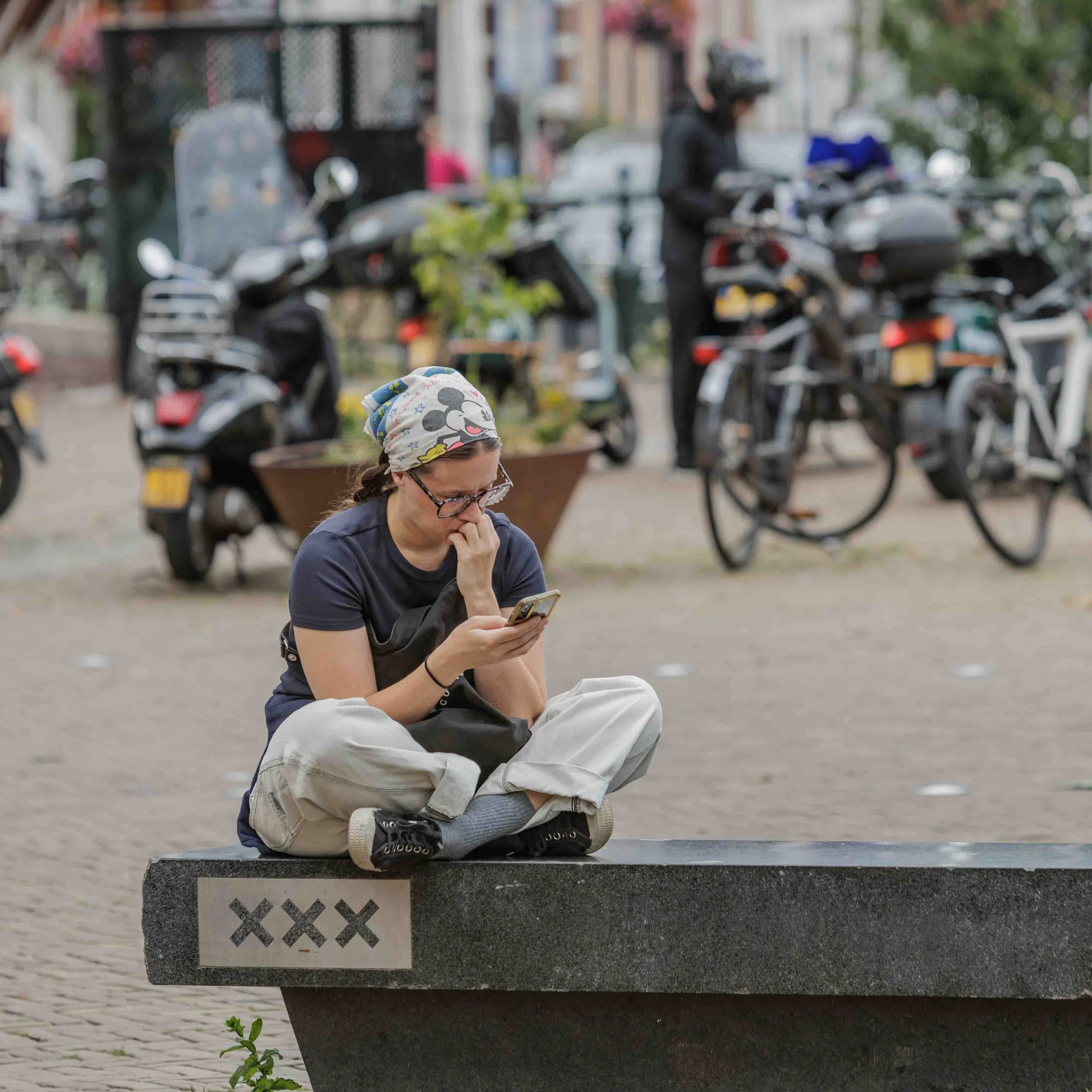 Vrouw zit op een bankje, kijkt op haar telefoon, met een pet met Mickey Mouse op haar hoofd, op de achtergrond staan verschillende gemotoriseerde voertuigen en een vrouw die een helm draagt.