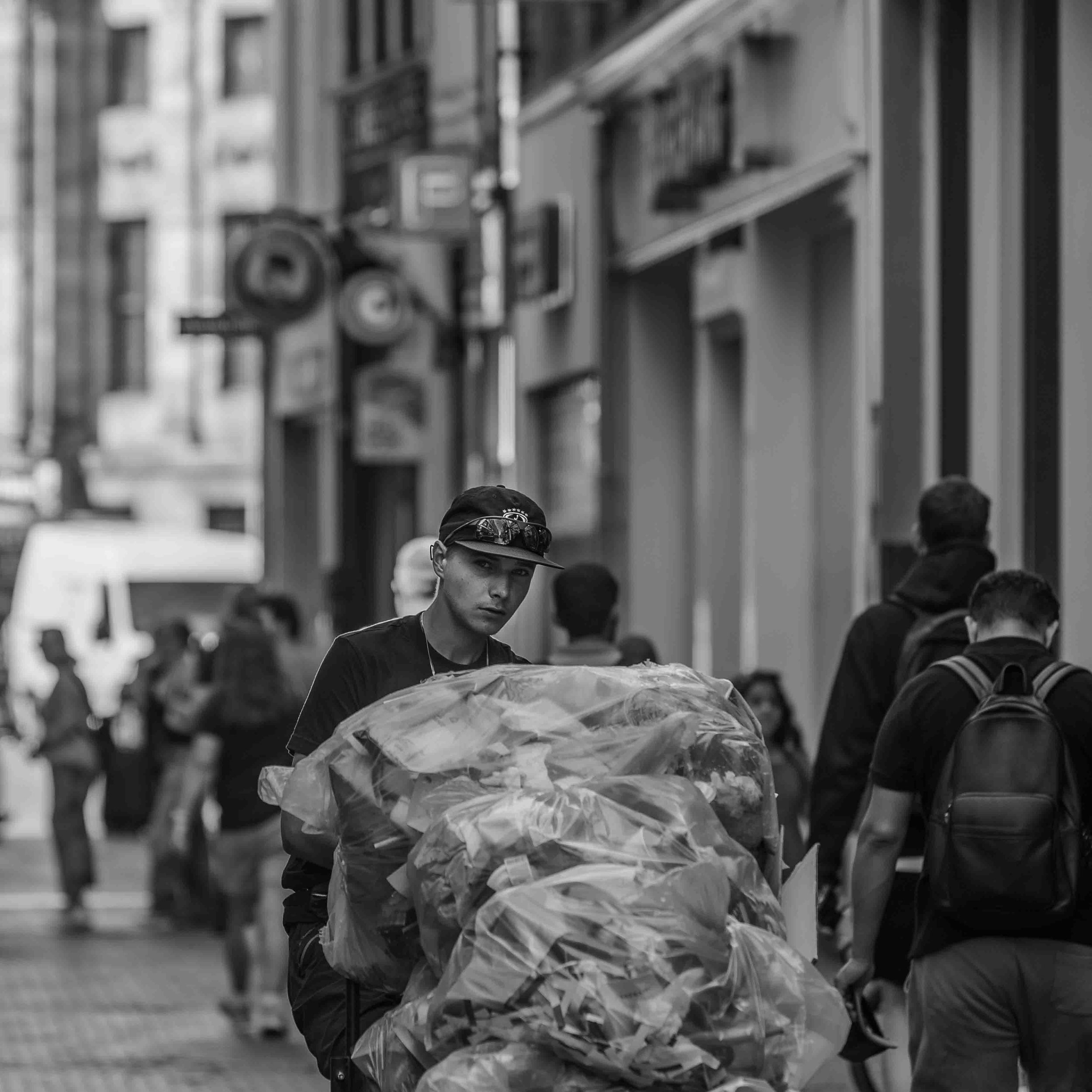 Een jongeman met een pet en een zwarte jas die een groot stuk plastic draagt in een drukke stadsstraat.