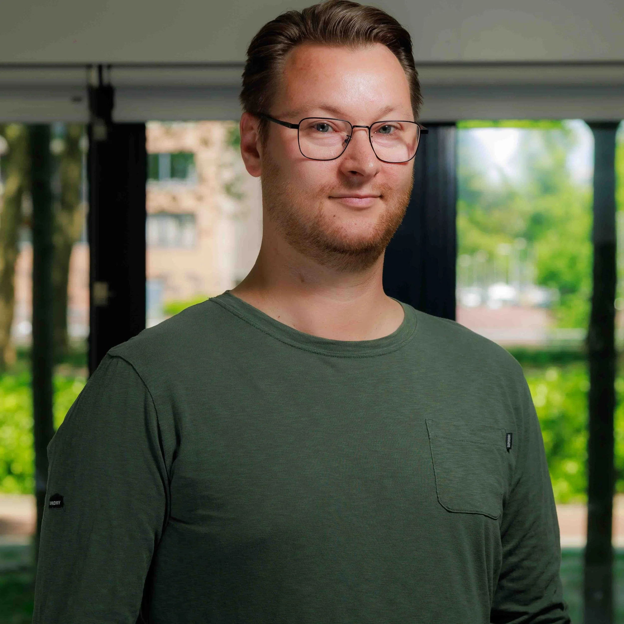 Close-up van een jonge man met een glimlach, draagt een groene t-shirt en bril, staat binnen met grote ramen en groene bomen buiten.