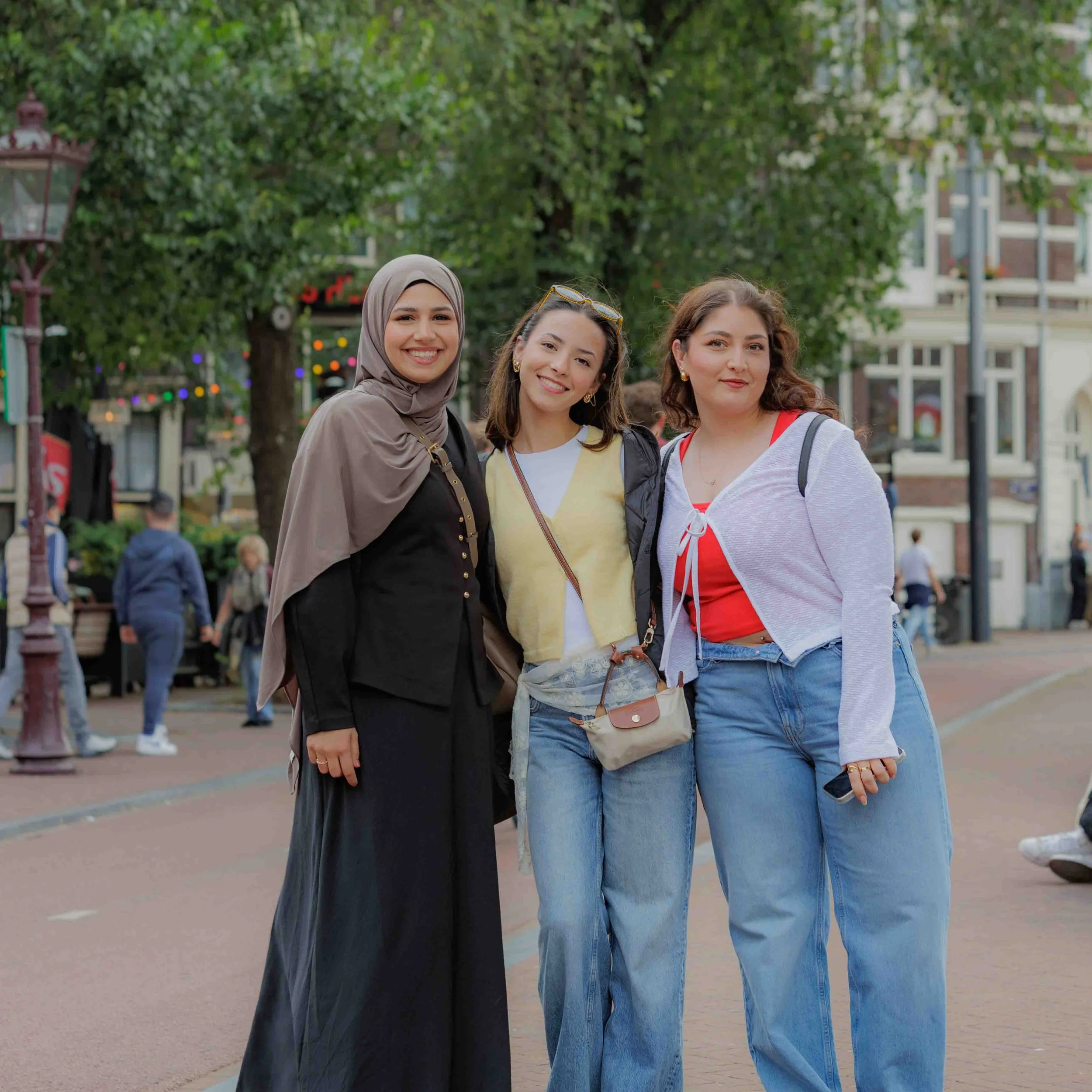 Drie jonge vrouwen lopen samen in een park met bomen en gebouwen op de achtergrond.