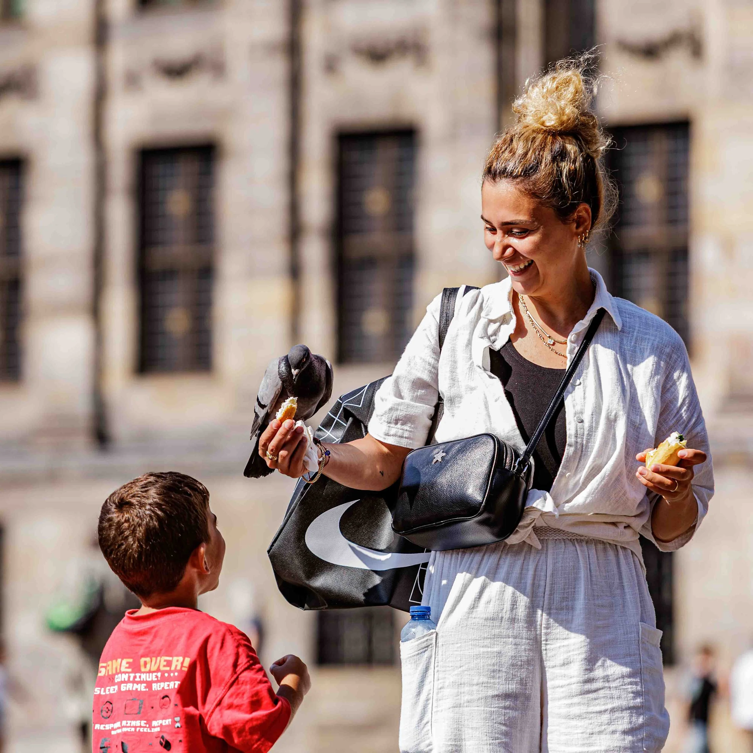 Vrouw met een lichte blouse en een zwart-witte tas over haar schouder, lachend en een ijsje vasthoudend, terwijl ze samen met een jongen in een rode shirt in een stadsomgeving staat.