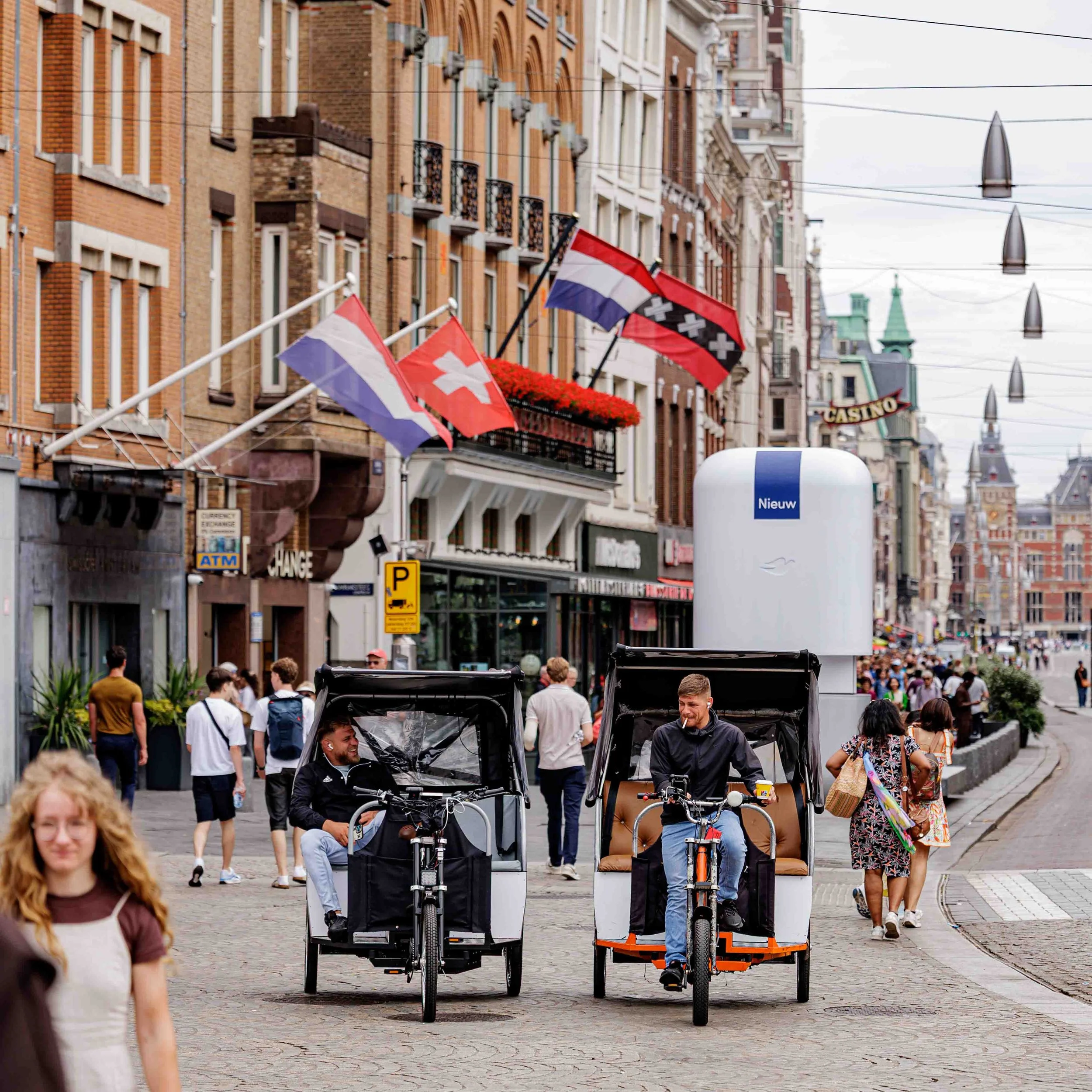 Straatbeeld met mensen, fietsen en vlaggen in een drukke stadsomgeving met historische gebouwen.