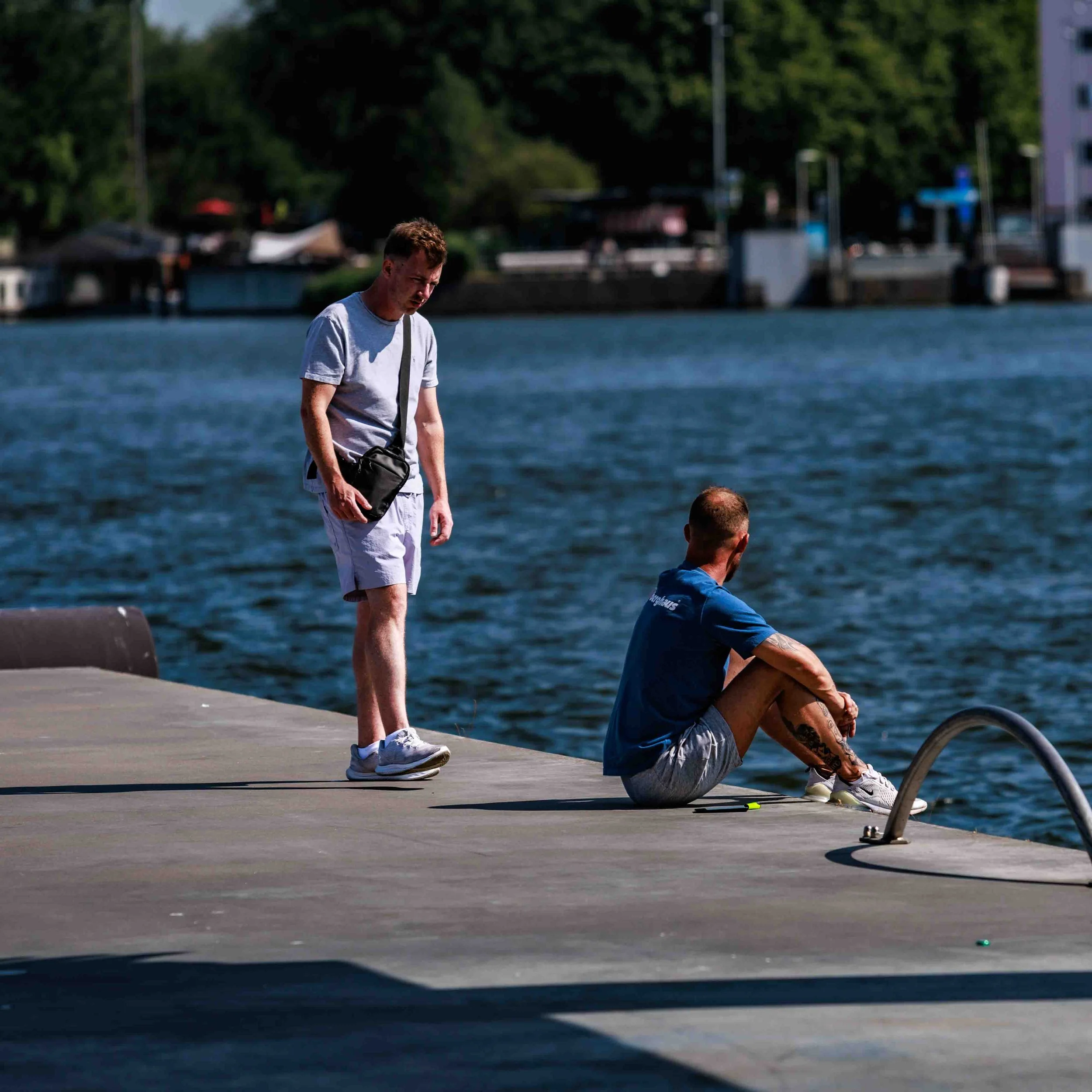 Een man in een grijs T-shirt en witte korte broek staat op een doorwaadbare plek aan een waterkant, terwijl een andere persoon op de grond voor hem zit.