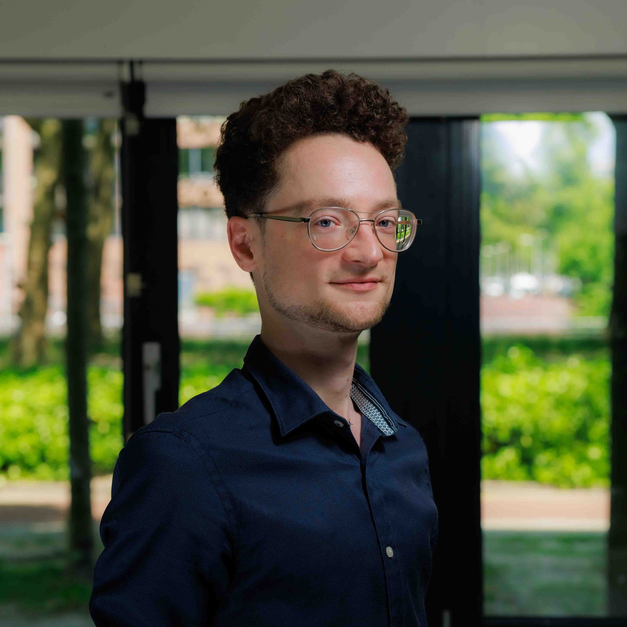 Portret van een jonge man met kort, krullend haar en bril, gekleed in een donkerblauw shirt, lachend op een groene achtergrond buiten.