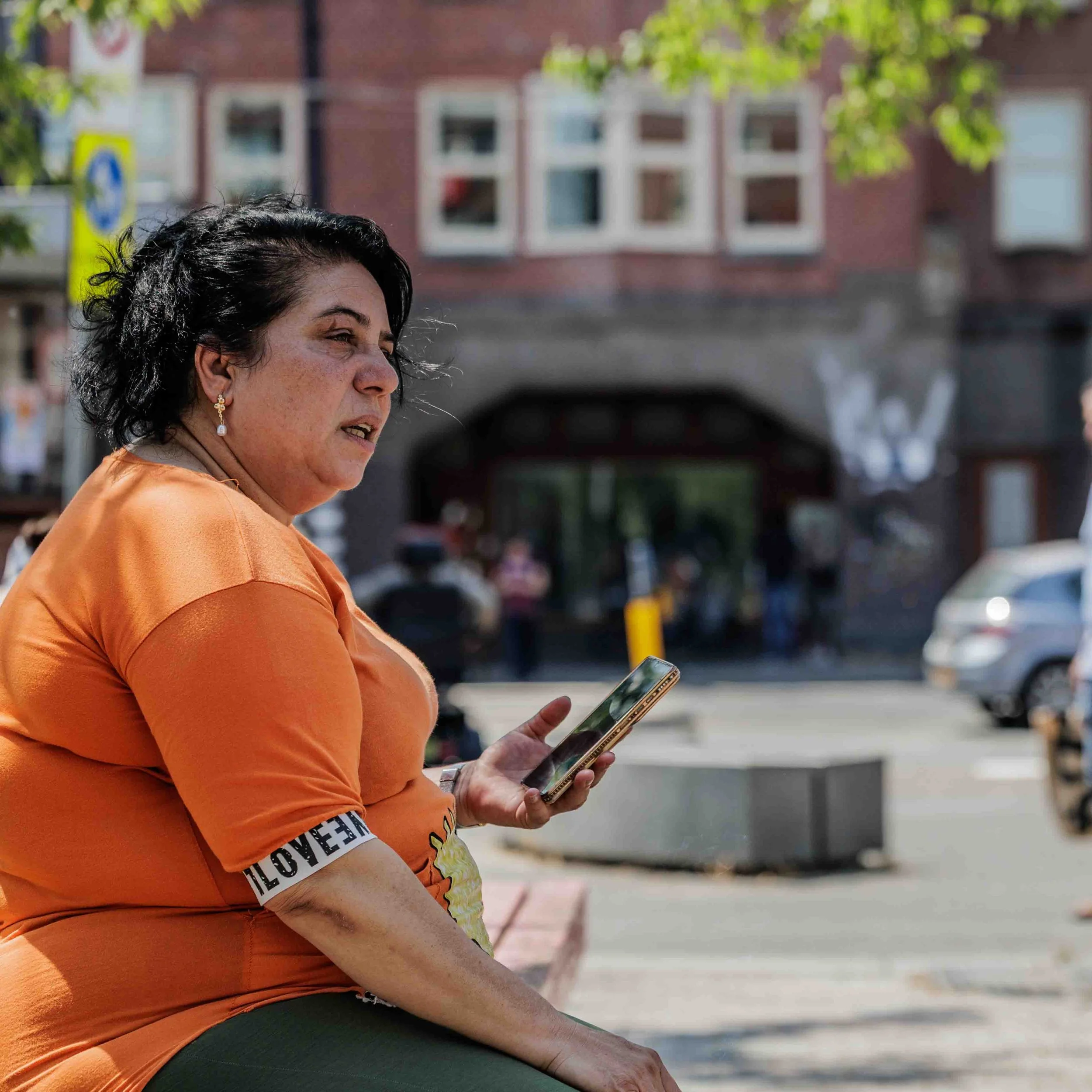 Vrouw met korte zwarte haar en oren met oorringen zit buiten op een bank, iemand gebruikt haar smartphone.