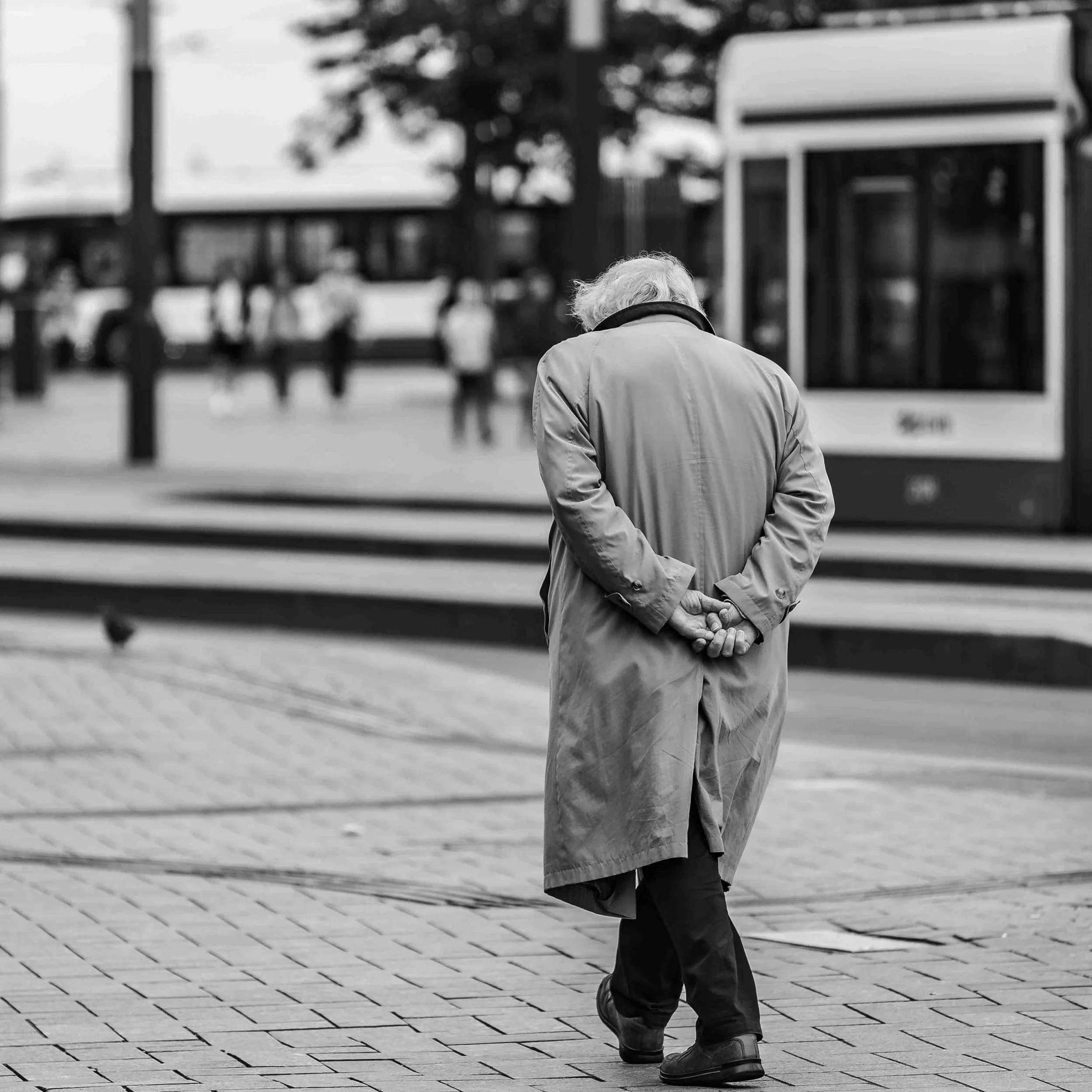 Een oudere persoon met grijs haar die een jas draagt, staat op een stenen bestrating en kijkt naar beneden met de handen achter de rug. Op de achtergrond is een bushokje en meerdere mensen die wachten.