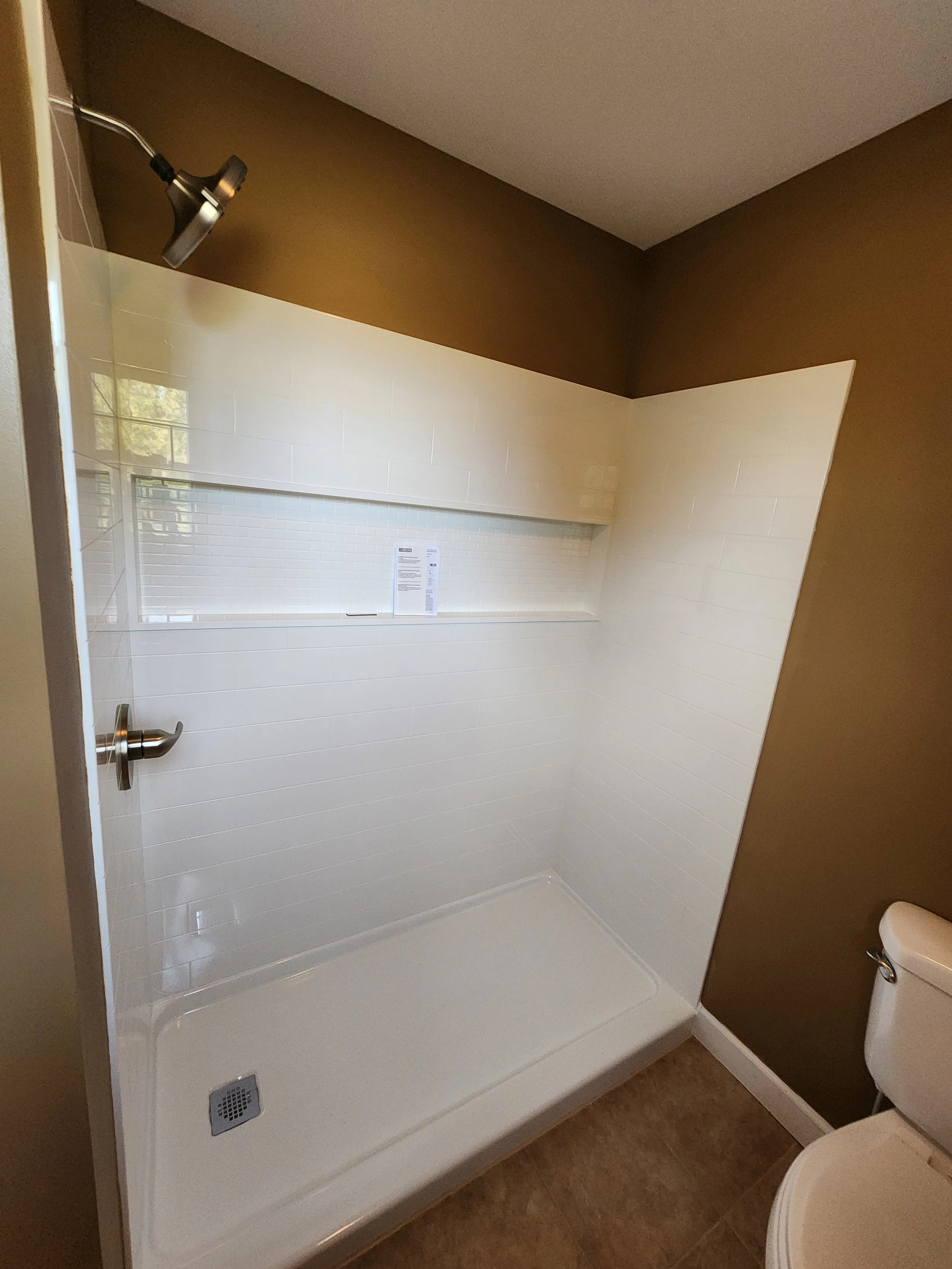 Bathroom with a prefabricated, cost-efficient shower, white tile walls, brown wall paint, a window, a toilet, and a shower drain on the floor.