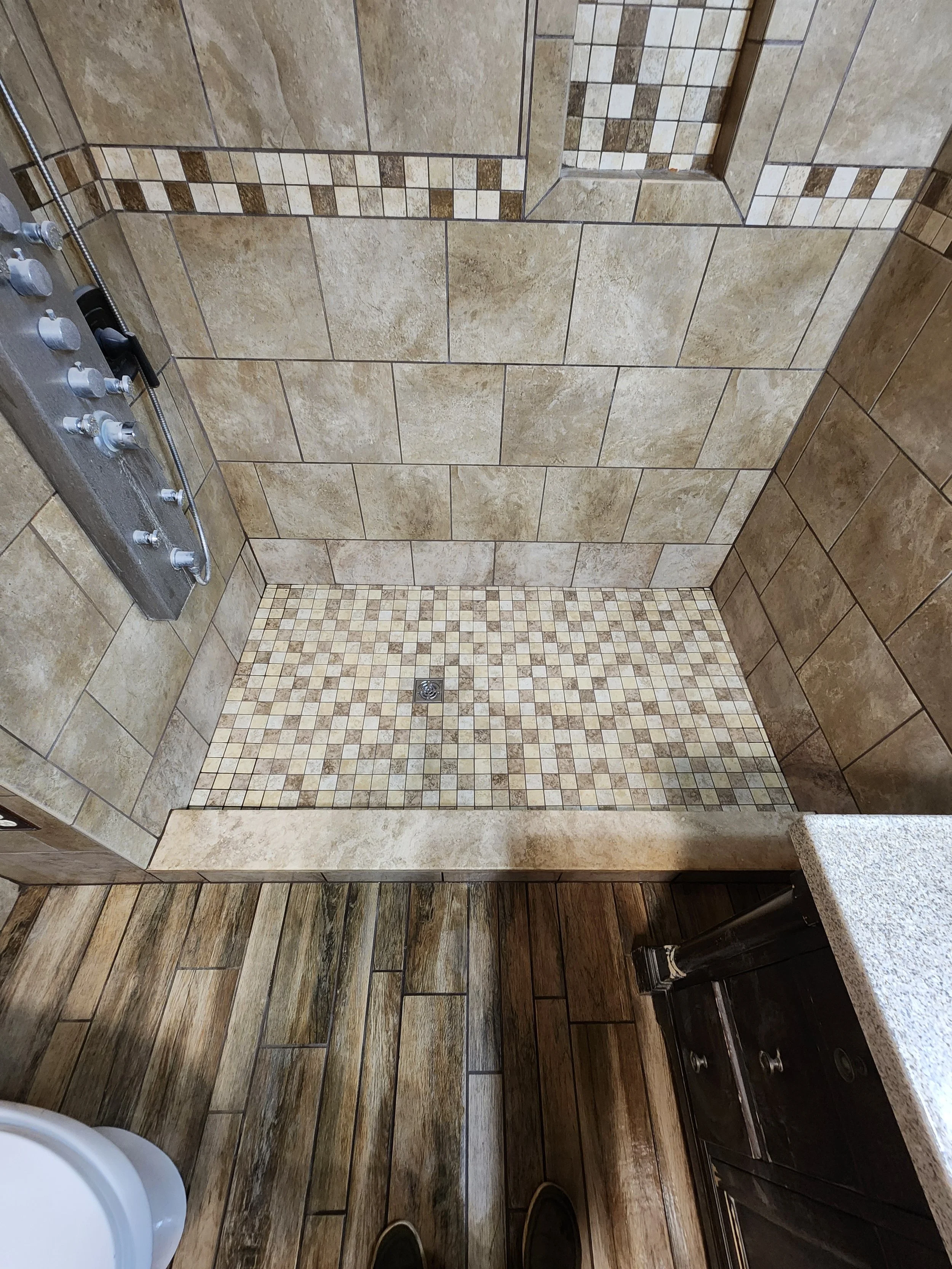 A tiled walk-in shower with large beige tiles on the walls, a decorative border of small square tiles, a built-in corner shelf, a drain on the shower floor, and a shower panel with multiple controls and jets.