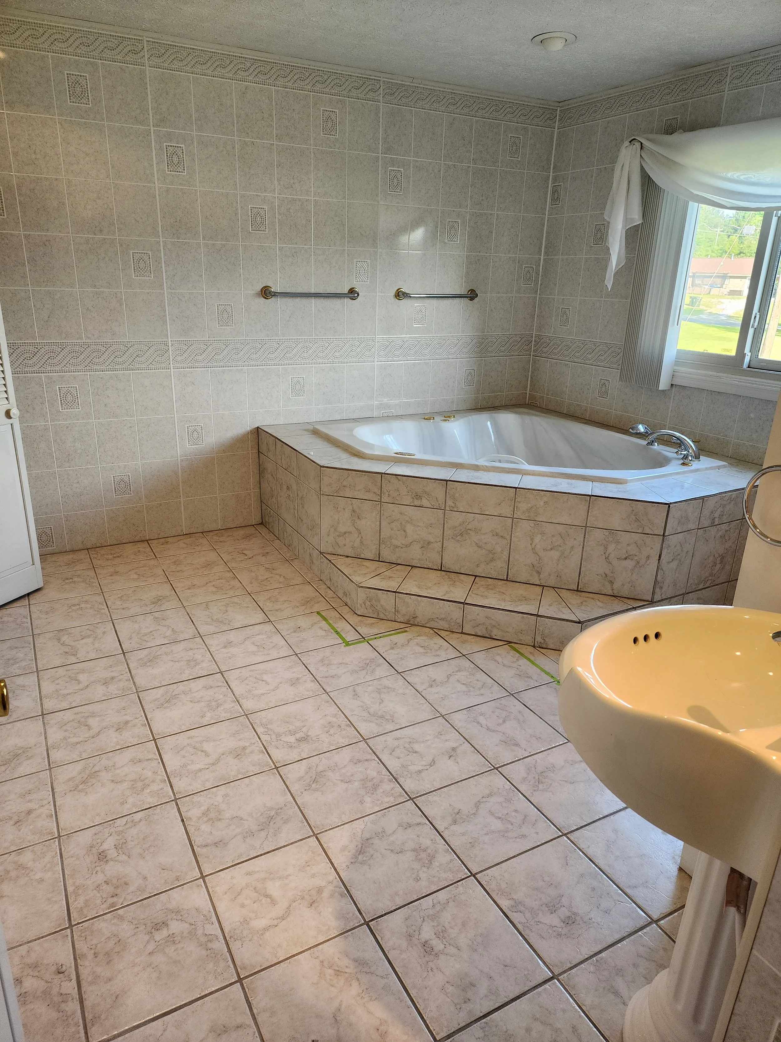 A bathroom with tiled floors and walls. There is a corner bathtub with a marble exterior, a window with a curtain, and a beige pedestal sink. Two towel bars are mounted on the wall above the bathtub.